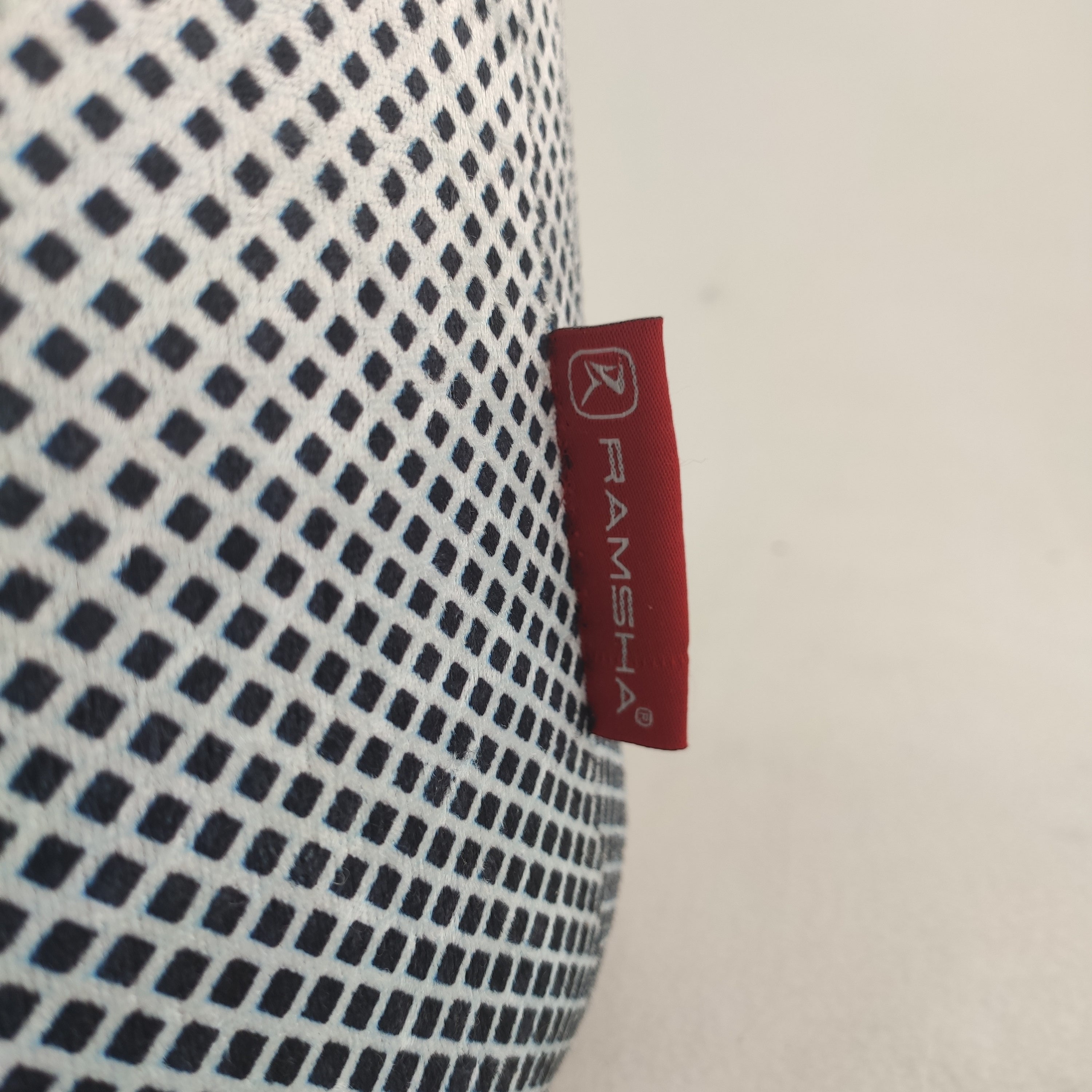 A close-up of the Printed Pedestal Pouf With Leather Handle PF-026 by RAMSHA HOME, featuring a black-and-white diamond fabric and a red RAMSHA tag with white logo, set against a plain light backdrop—ideal for modern home decor.