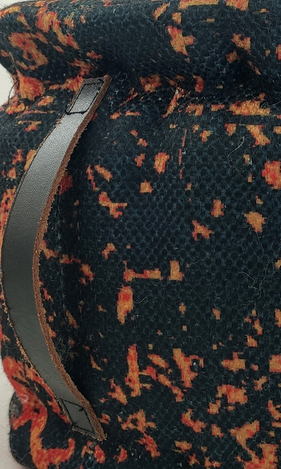 Close-up of the RAMSHA HOME Printed Multi Color Pouf With Leather Handle, showcasing textured polyester fabric with orange, yellow, and red speckles on a black background plus a dark leather handle on the left.