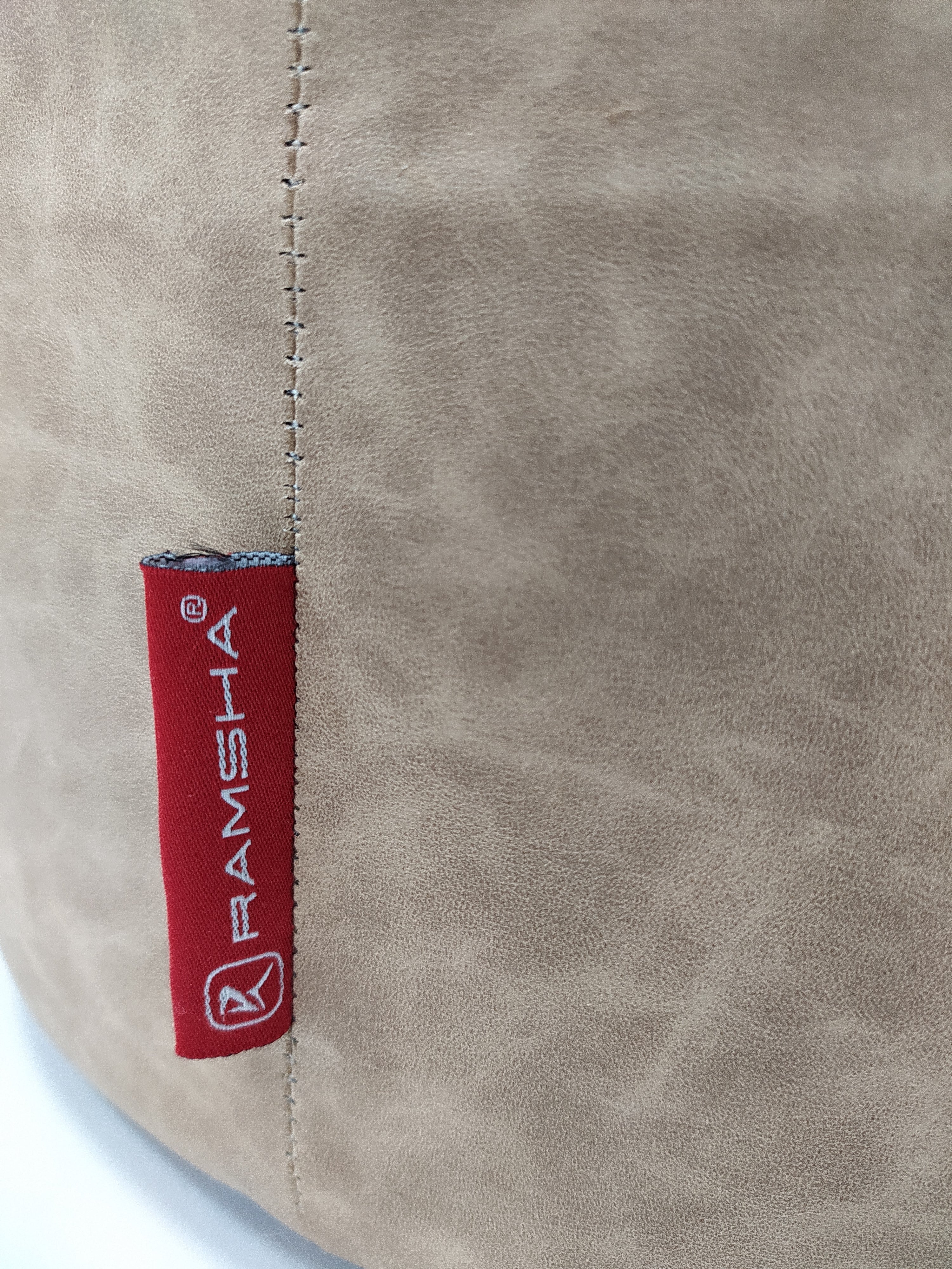 Close-up of a beige Leather Cylinderical Pouf from RAMSHA HOME, featuring visible stitching and a red fabric tag with the brand name and circular logo sewn onto this stylish seating accent.