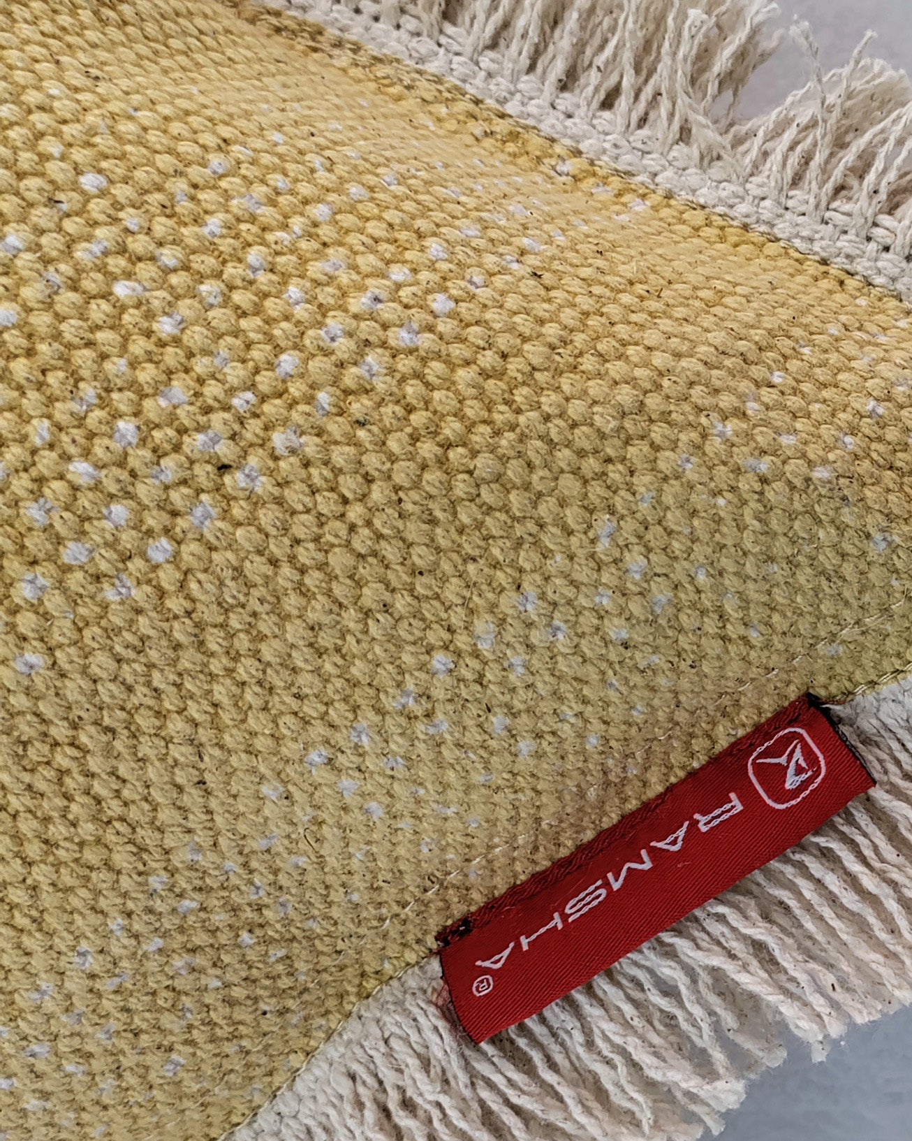 Close-up of the Cotton Cushion With Fringes cs-141 by RAMSHA HOME, featuring yellow cotton fabric with white speckles, beige fringe edges, and a red RAMSHA HOME tag on one corner.