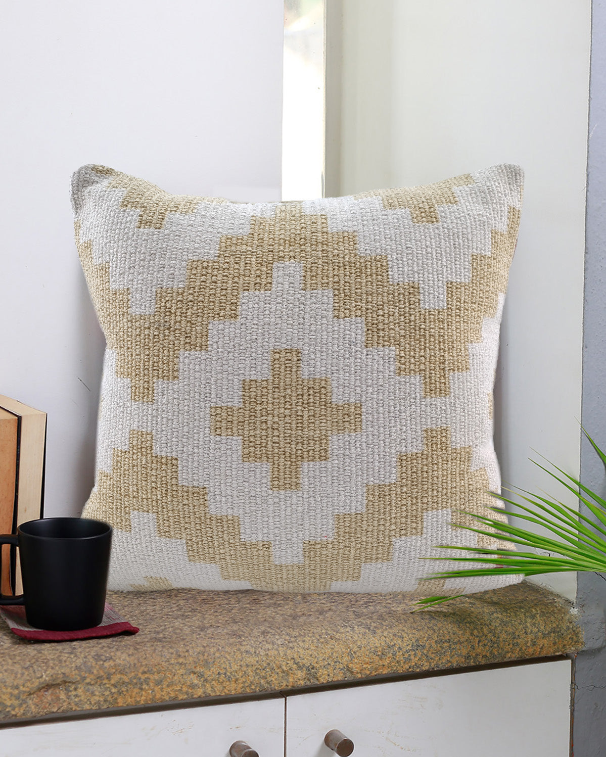 The RAMSHA HOME Zigzag Design Pattern Cotton Cushion CS-179, with its beige and white tones, sits on a stone surface near a window. A black mug on a red coaster, a book, and a green plant complement this cozy setup.