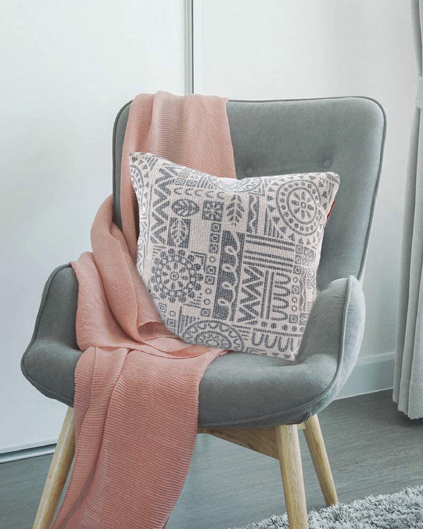 A gray cushioned chair with wooden legs features the RAMSHA HOME Grey and White Color Cotton Cushion CS-182 and a soft pink throw, set on a wooden floor near a window with white curtains.