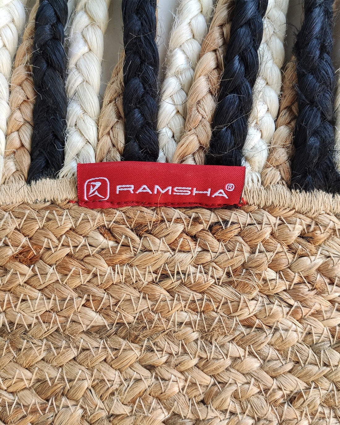 Close-up of the Amazing Handmade Braided Round Jute Rug - BR 001 by RAMSHA HOME, featuring alternating black, white, and natural-fiber braids with a red fabric label and logo attached to this stylish round jute rug.