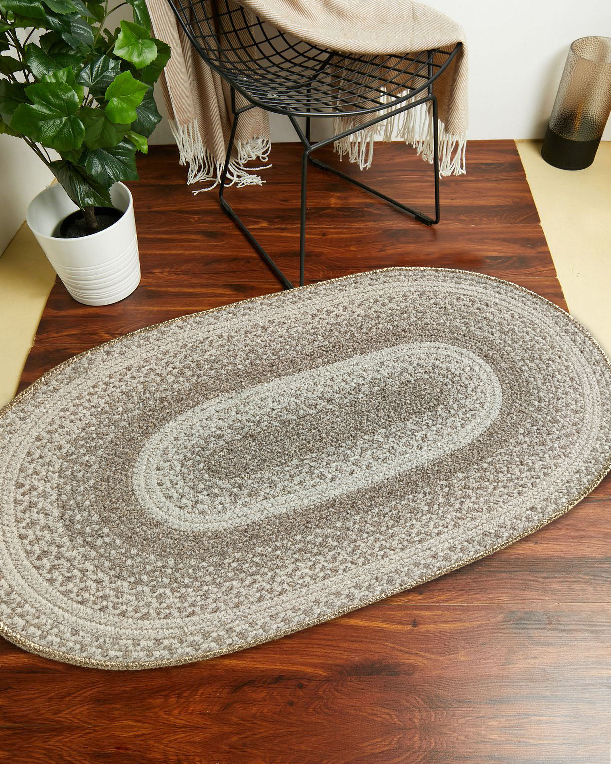 The Luxurious Handmade Multi Color Wool Floor Braided Rug - BR 044 by RAMSHA HOME rests on a wooden floor, near a black chair with a beige fringed throw, a potted plant, and a decorative lantern.