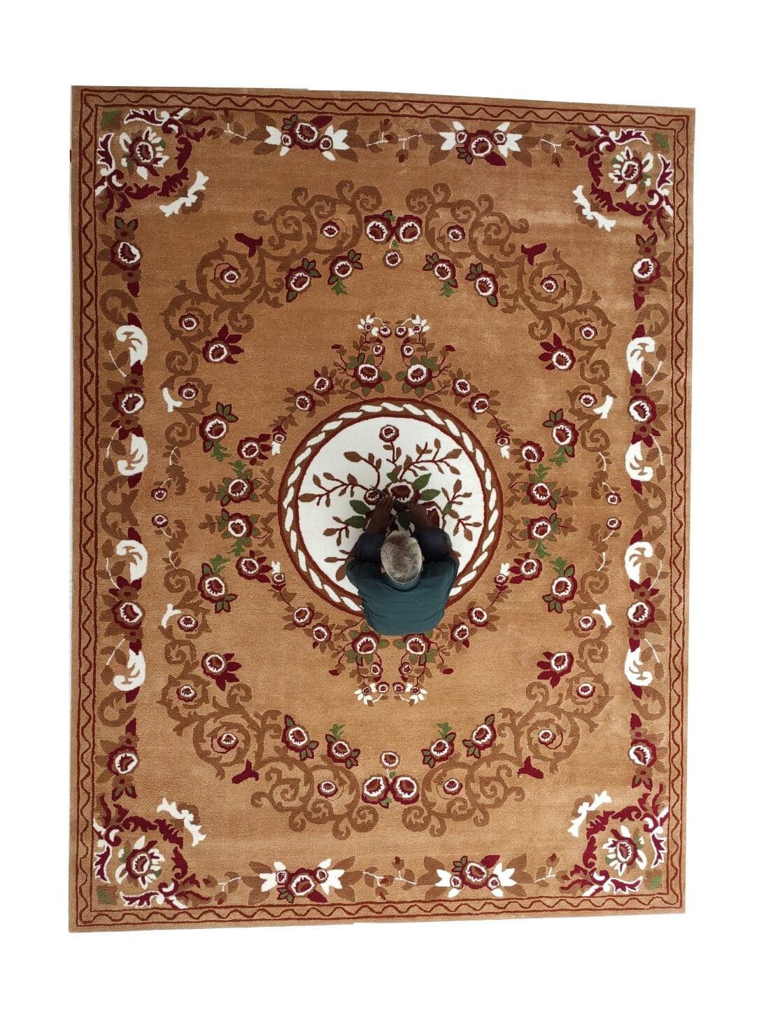 A person in dark clothes sits cross-legged on a RAMSHA HOME Amazing Polyester & Cotton Hall Decor Rug - HTP 06 (300x400cm), featuring a tan ornate medallion with red, green, and white floral patterns, viewed from above.