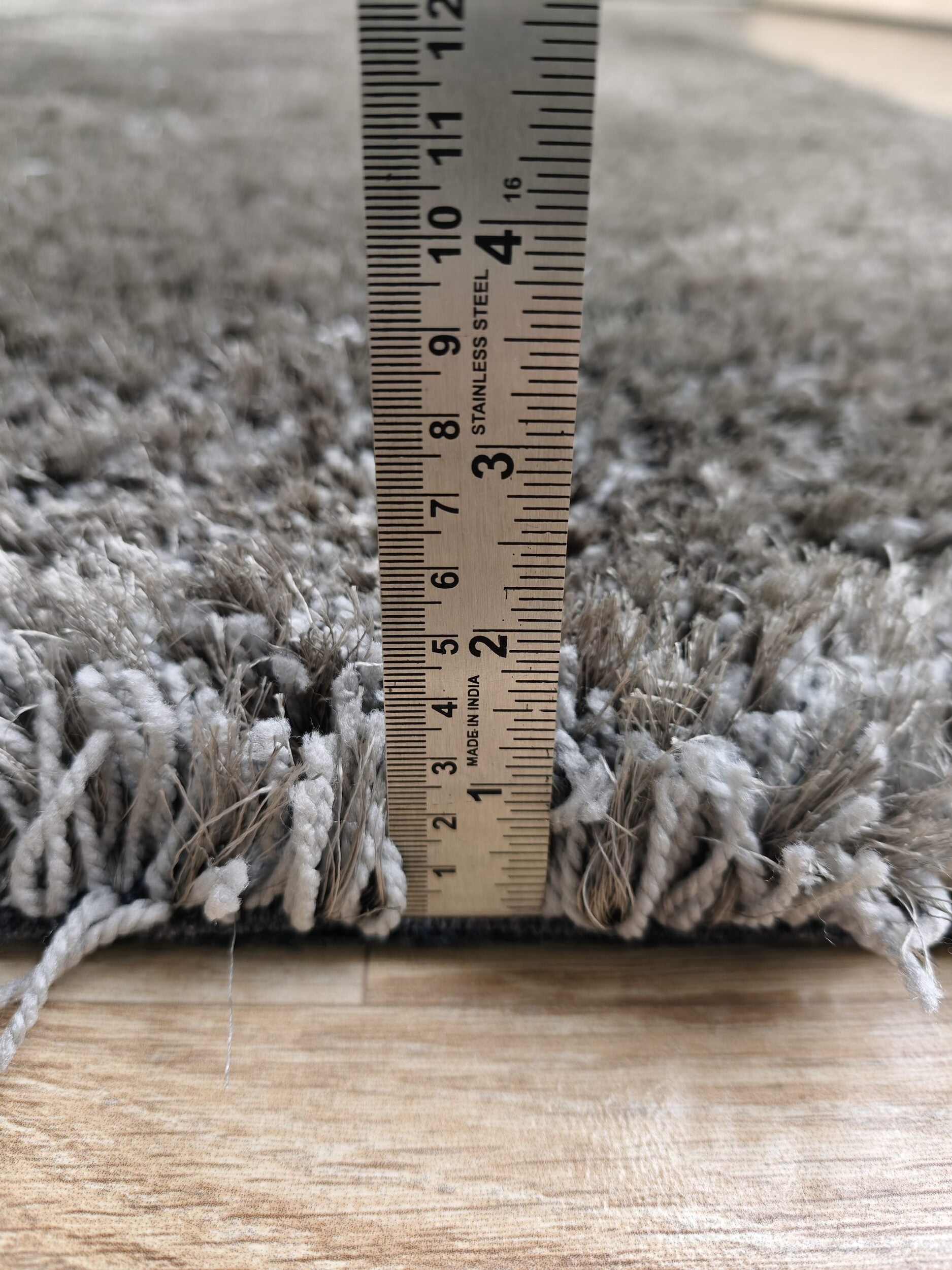 A ruler stands vertically on a wooden floor, measuring the height of the RAMSHA HOME Grey Color Rectangle Shaped Shaggy Rug - SG 05. The rug’s pile measures approximately 1 inch thick.