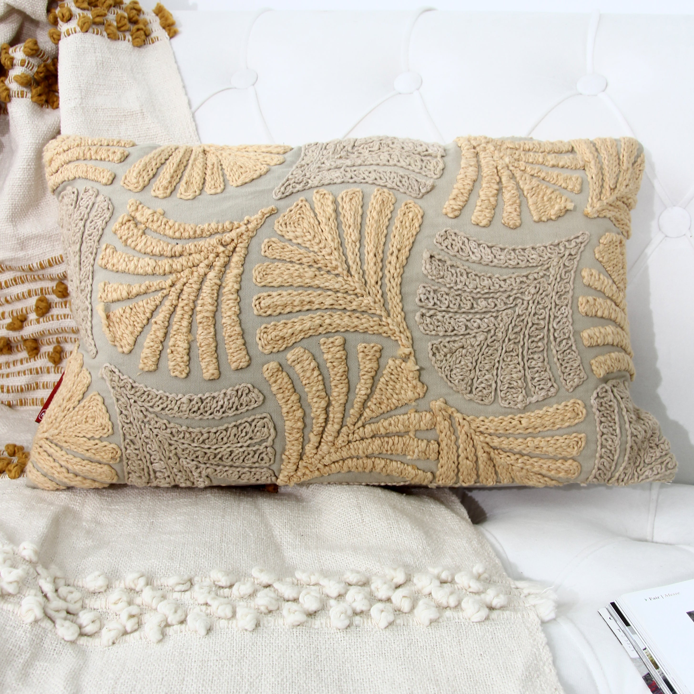 The RAMSHA HOME Multi Color Embroidery Cotton Cushion CS-263, featuring textured leaf-like patterns in tan and light brown, rests on a white couch alongside cream-colored, textured throws.