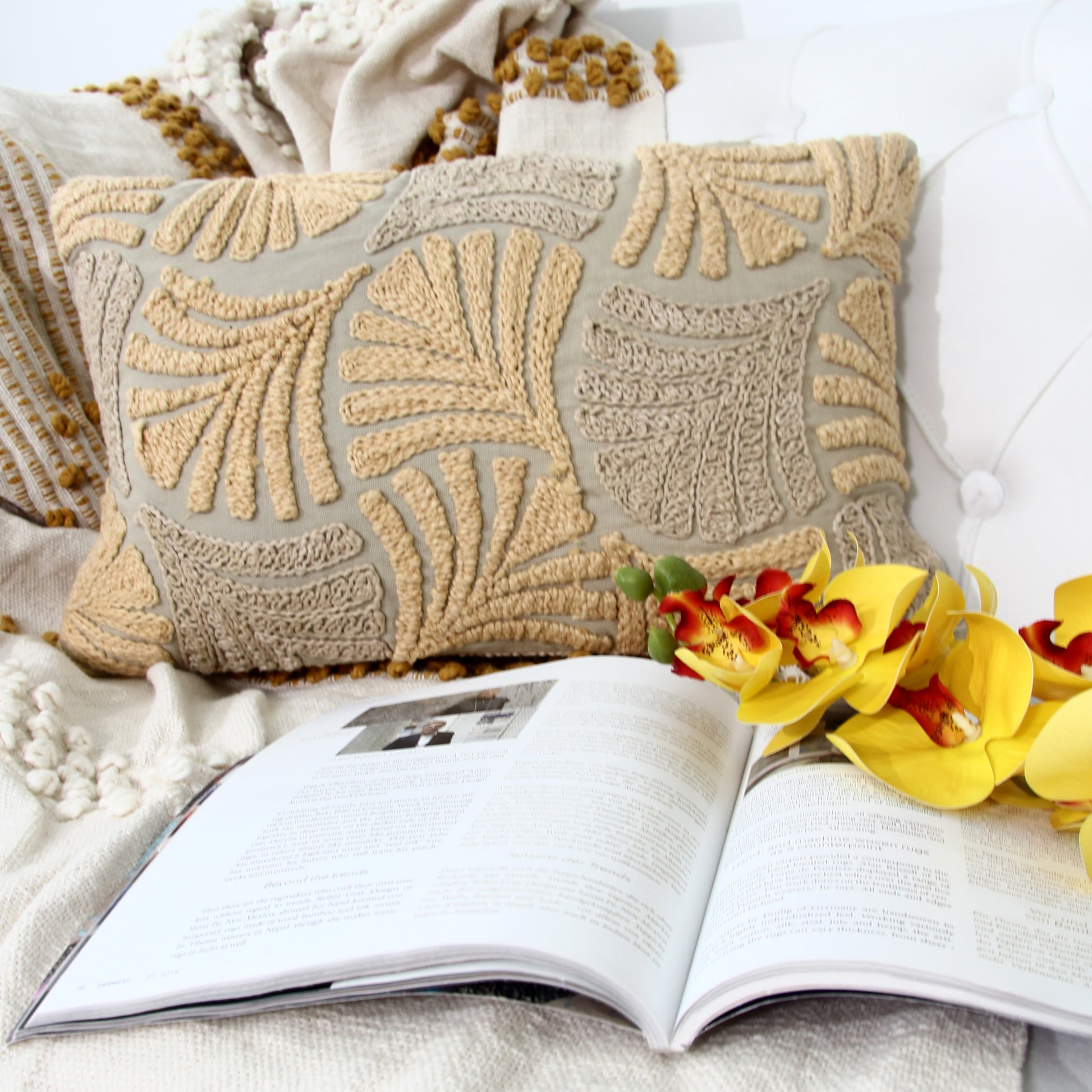The RAMSHA HOME Multi Color Embroidery Cotton Cushion CS-263 rests on a cream knit blanket with an open book and yellow flowers in front, creating a cozy reading nook with this stylish decorative pillow.