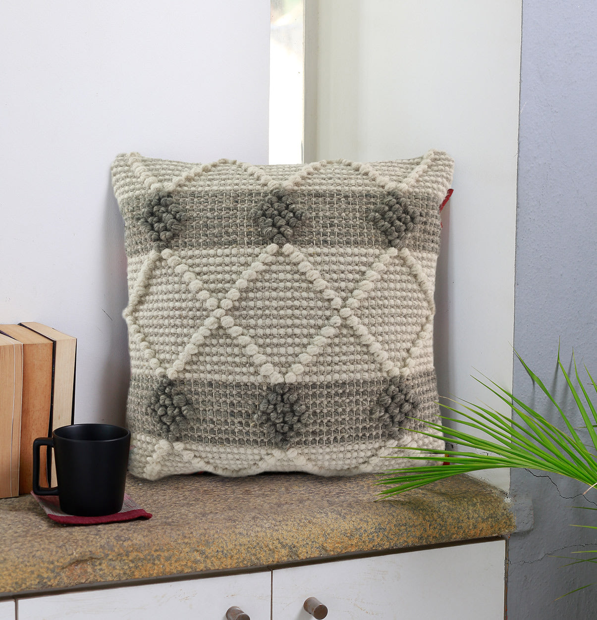 The Embroidery Woven Cushion With Insert cs-162 by RAMSHA HOME, in cream and gray with a geometric pattern, rests on a countertop near a window beside a black mug, stacked books, and a green plant.