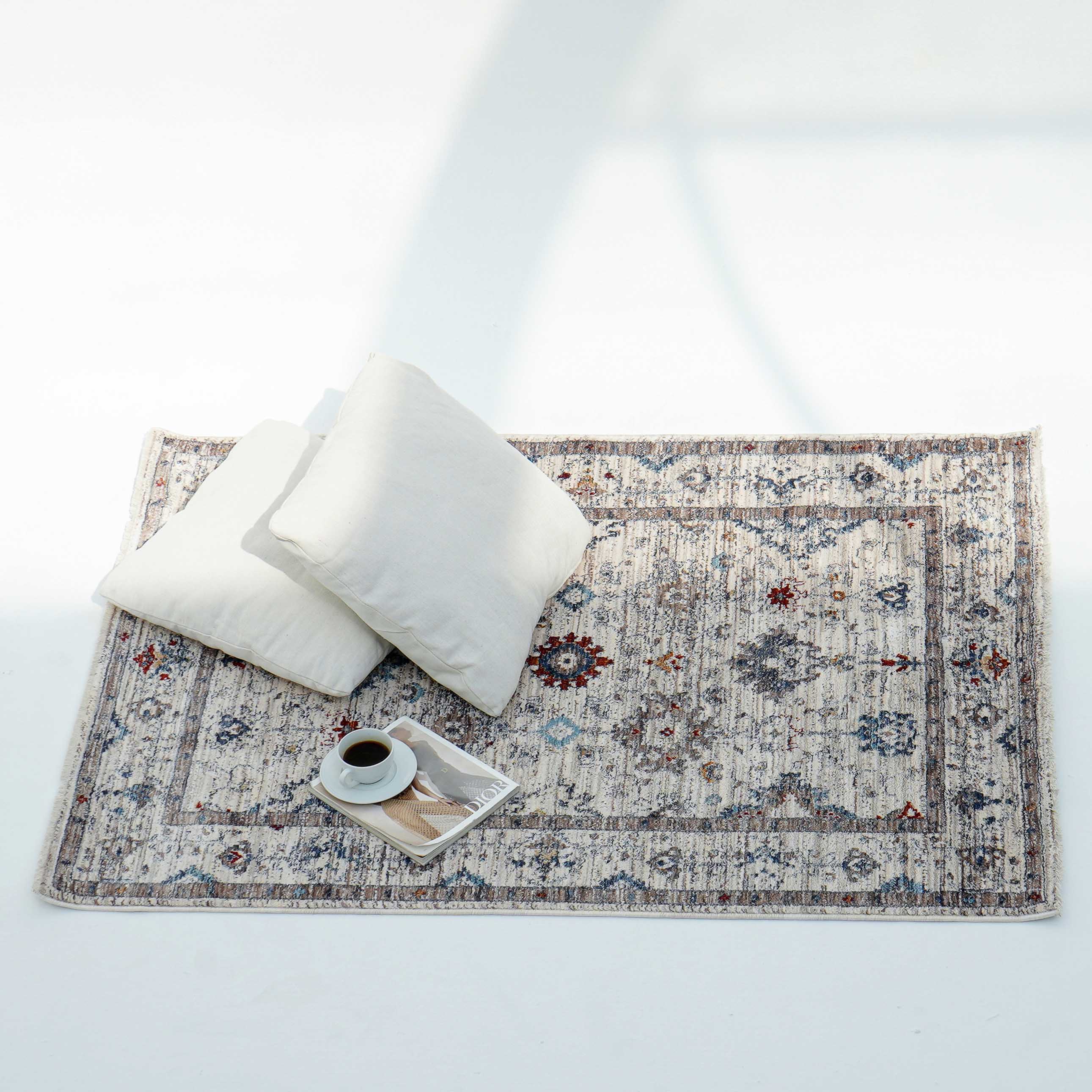 Two white pillows and a tray with a coffee cup and book rest on the RAMSHA HOME Cream Color Indoor Area Rug K655I, its geometric pattern in blue, red, and beige tones accenting the bright white floor.