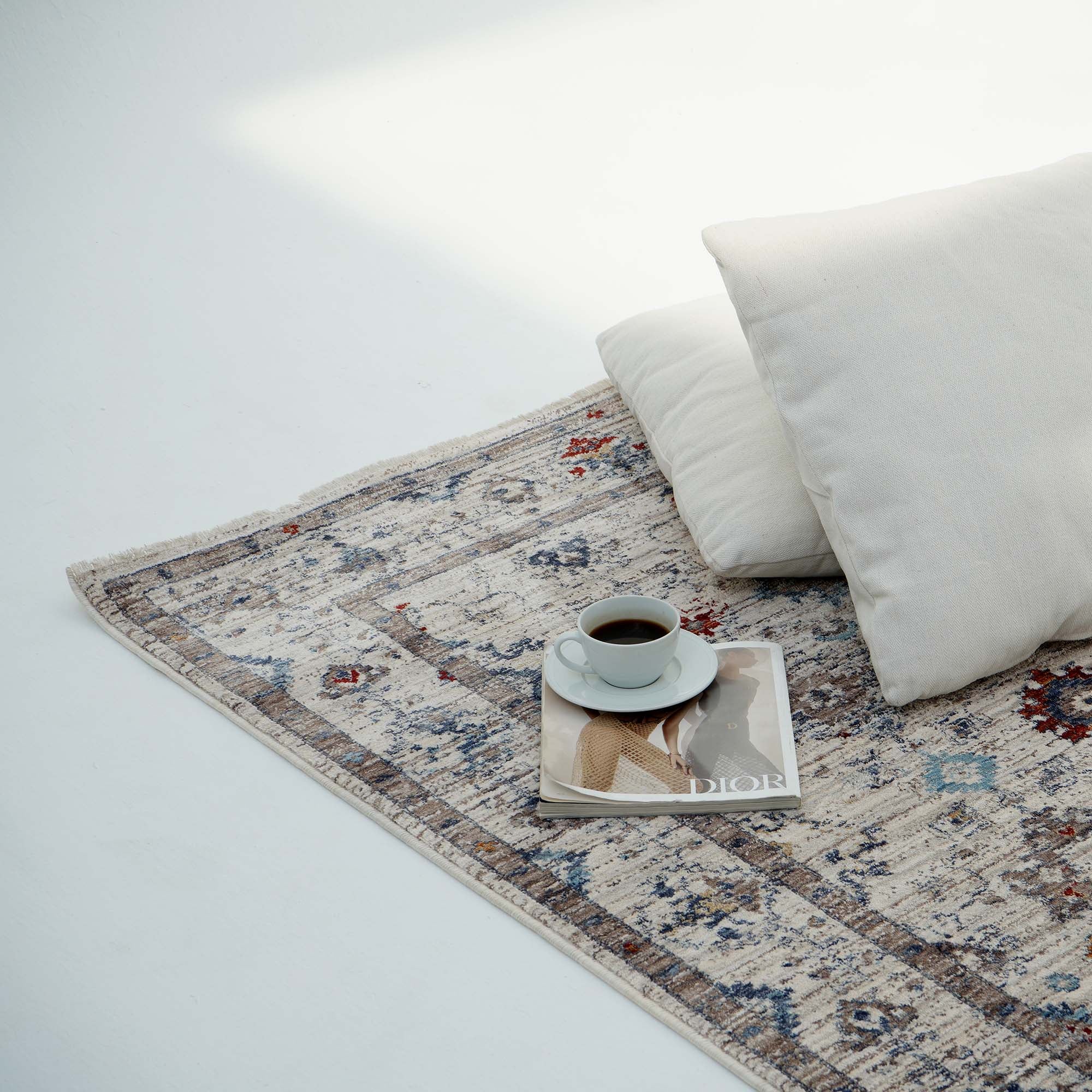 A cup of coffee on a saucer sits on a fashion magazine atop the RAMSHA HOME Cream Color Indoor Area Rug K655I, beside two white pillows in gentle natural light.