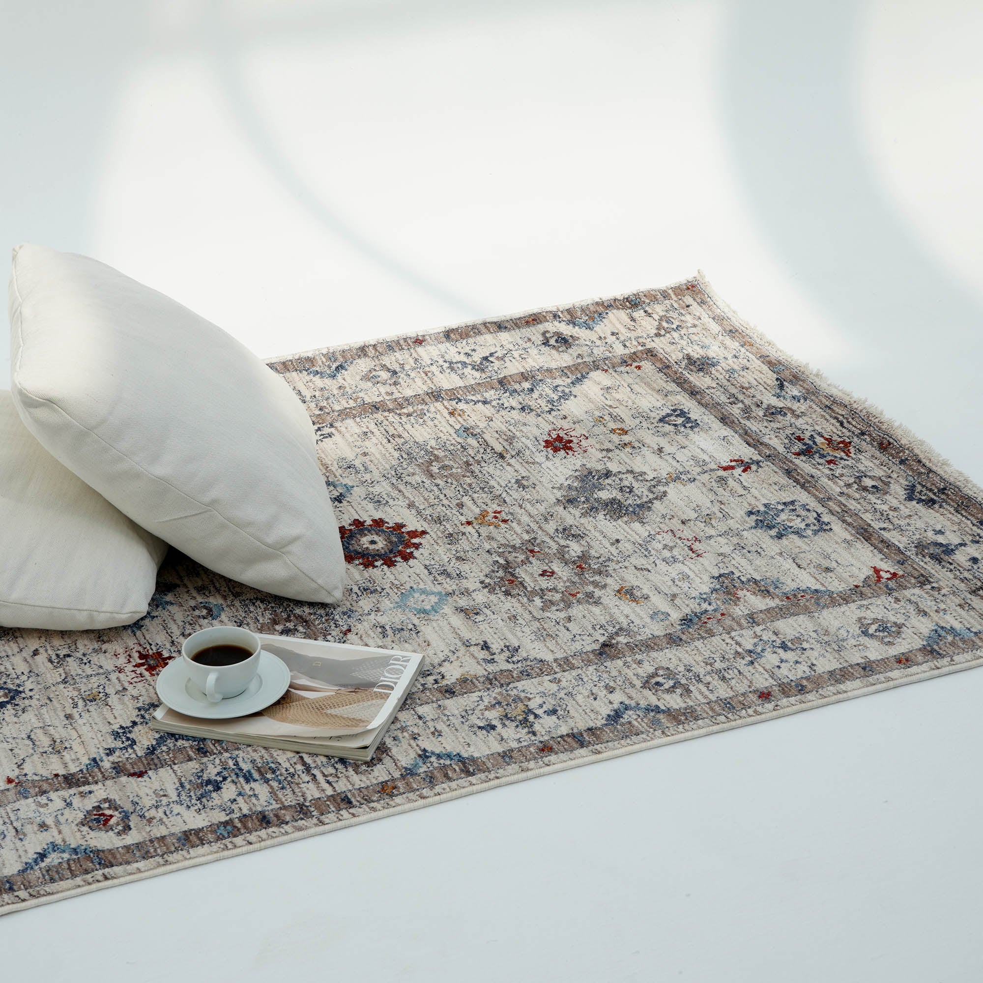 The Cream Color Indoor Area Rug K655I by RAMSHA HOME lies on a white floor, topped with two white pillows. A coffee cup and magazine rest on the rug, creating a cozy, inviting scene in gentle natural light.