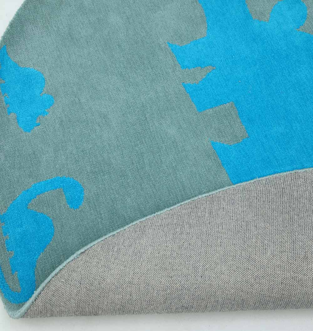 A close-up of the RAMSHA HOME Kids Dinosaur Rug - Blue & Green Color Playroom Area Carpet (KD 008, 150 cm) shows a corner flipped to reveal its grey backing, with blue and green dinosaur silhouettes on a grey background.