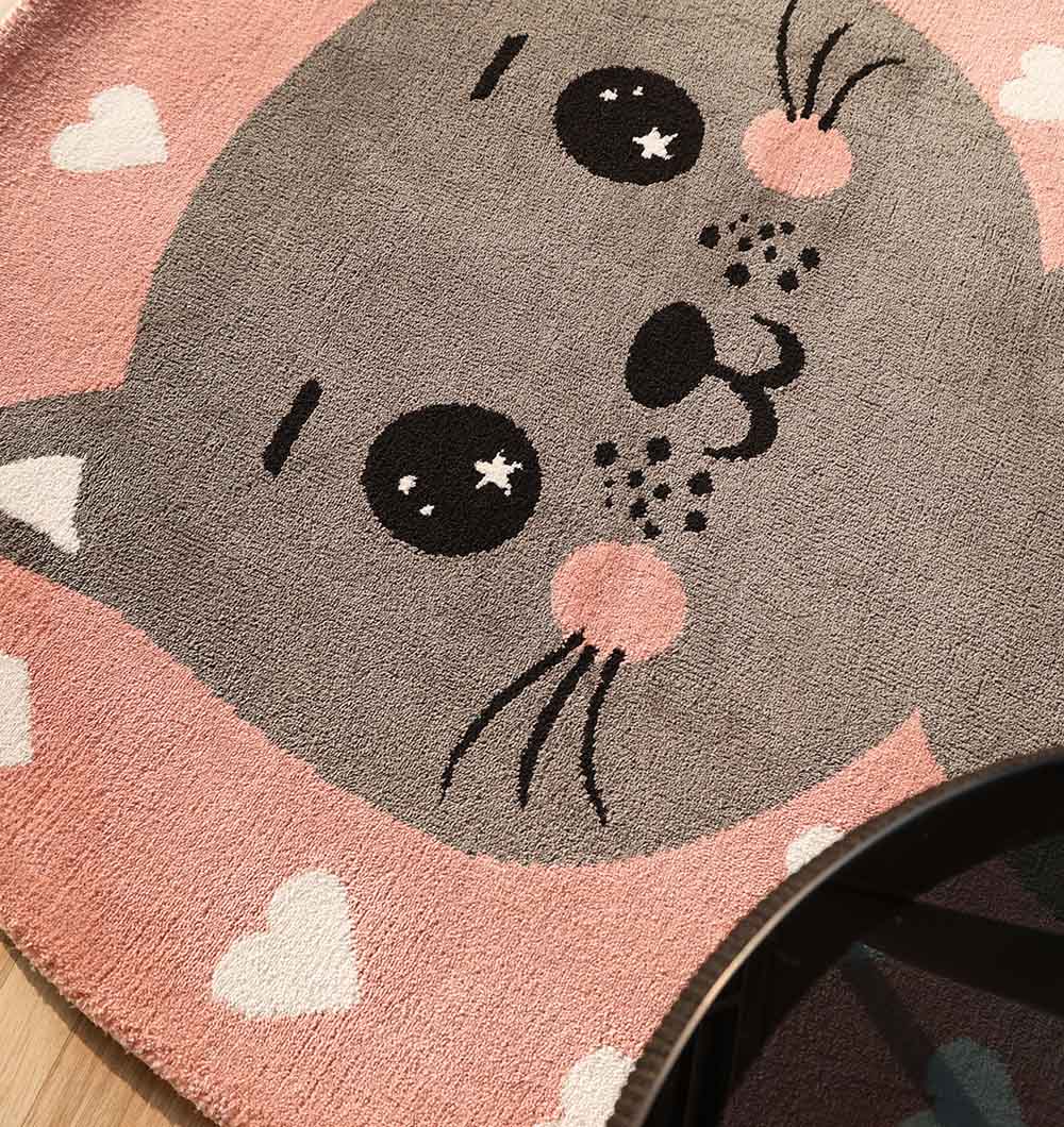 A close-up of the RAMSHA HOME Kids Cat Rug - Pink & Grey Color Playroom Area Carpet (KD 009, 120 cm) shows a cute cartoon cat face with blush cheeks, whiskers, big eyes, and white hearts on the edges. A glass table is partly visible in the corner.
