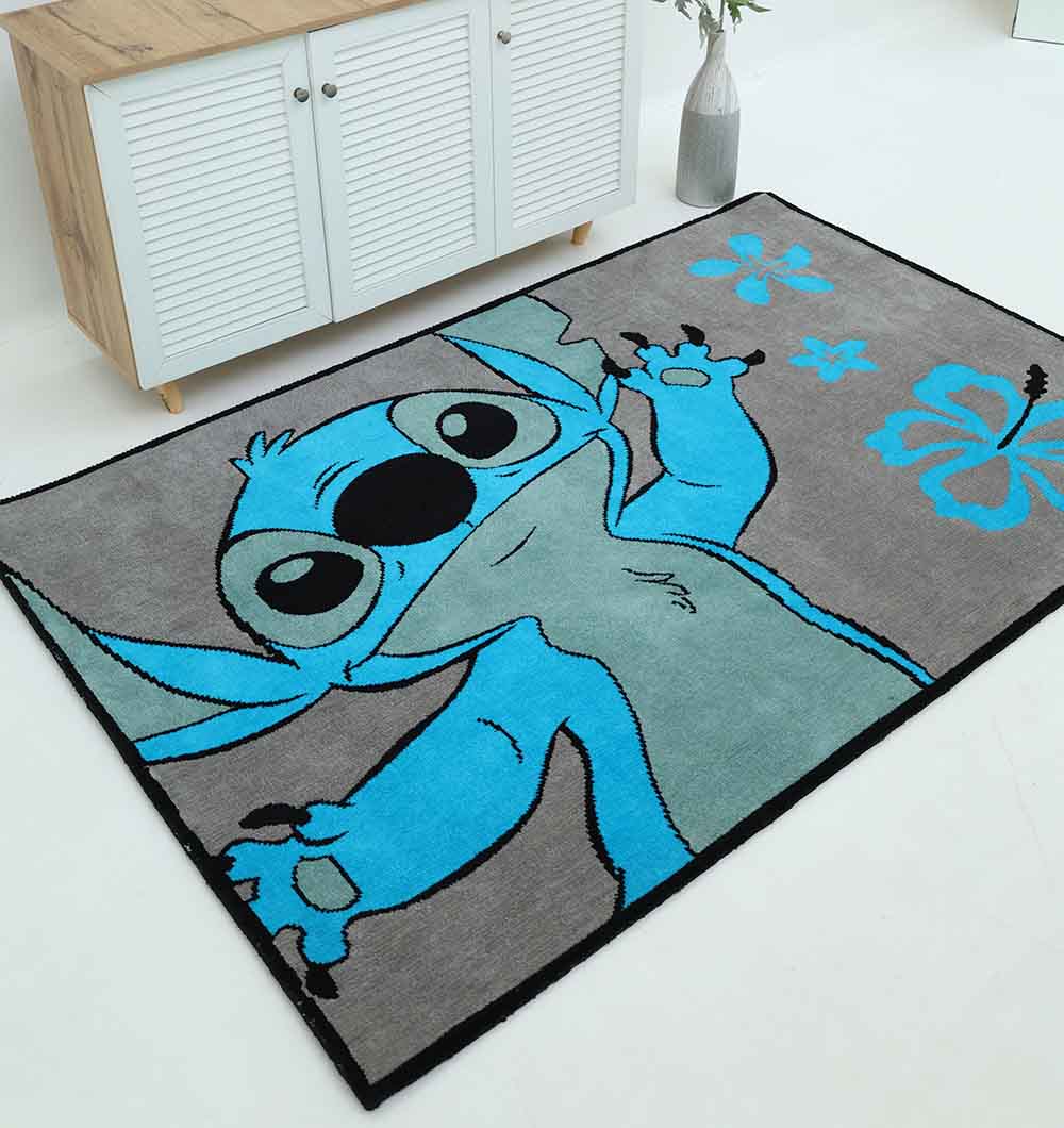 The RAMSHA HOME Kids Rug - Multi Color Playroom Area Carpet KD 010 (120x180 cm) features a cartoon blue character with big ears and flowers, perfect for playrooms and stylishly placed near a white cabinet and vase with greenery.