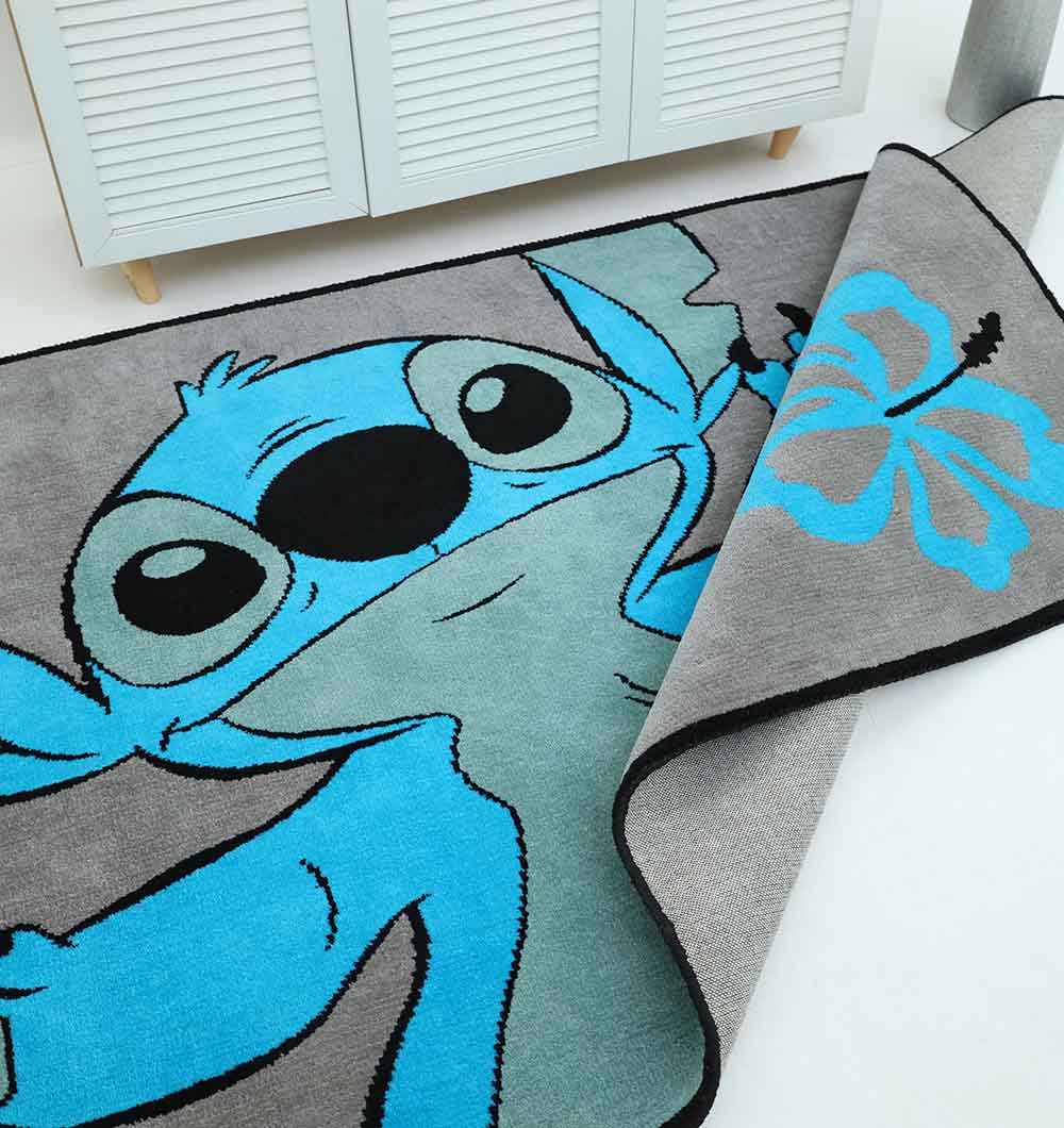 The RAMSHA HOME Kids Rug - Multi Color Playroom Area Carpet (KD 010, 120x180 cm) features a large cartoon Stitch design in blue and gray with a blue hibiscus flower, shown partially folded on a white floor near a cabinet.
