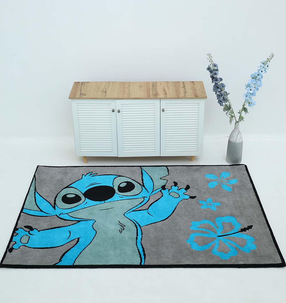 The RAMSHA HOME Kids Rug - Multi Color Playroom Area Carpet (KD 010, 120x180 cm) with a blue cartoon character and flowers is on a white floor before a white cabinet with wooden top; beside it stands a tall vase of purple flowers.