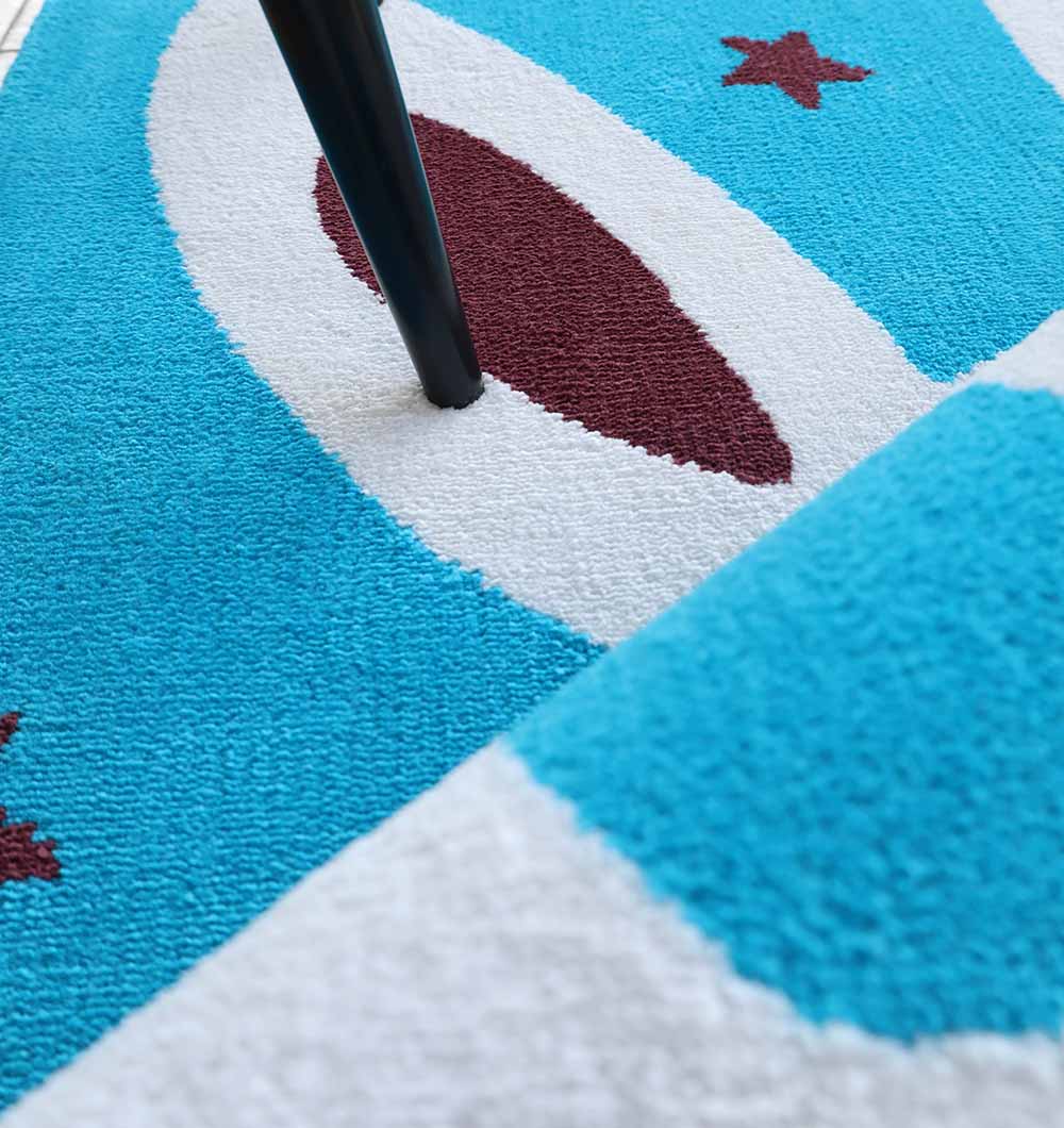 Close-up of the RAMSHA HOME Multi Color Kids Area Carpet - KD 011 (120x180 cm), featuring a blue, white, and brown abstract oval and star pattern. A black chair leg rests on this durable, kid-friendly polyester rug, with a folded edge in the foreground.
