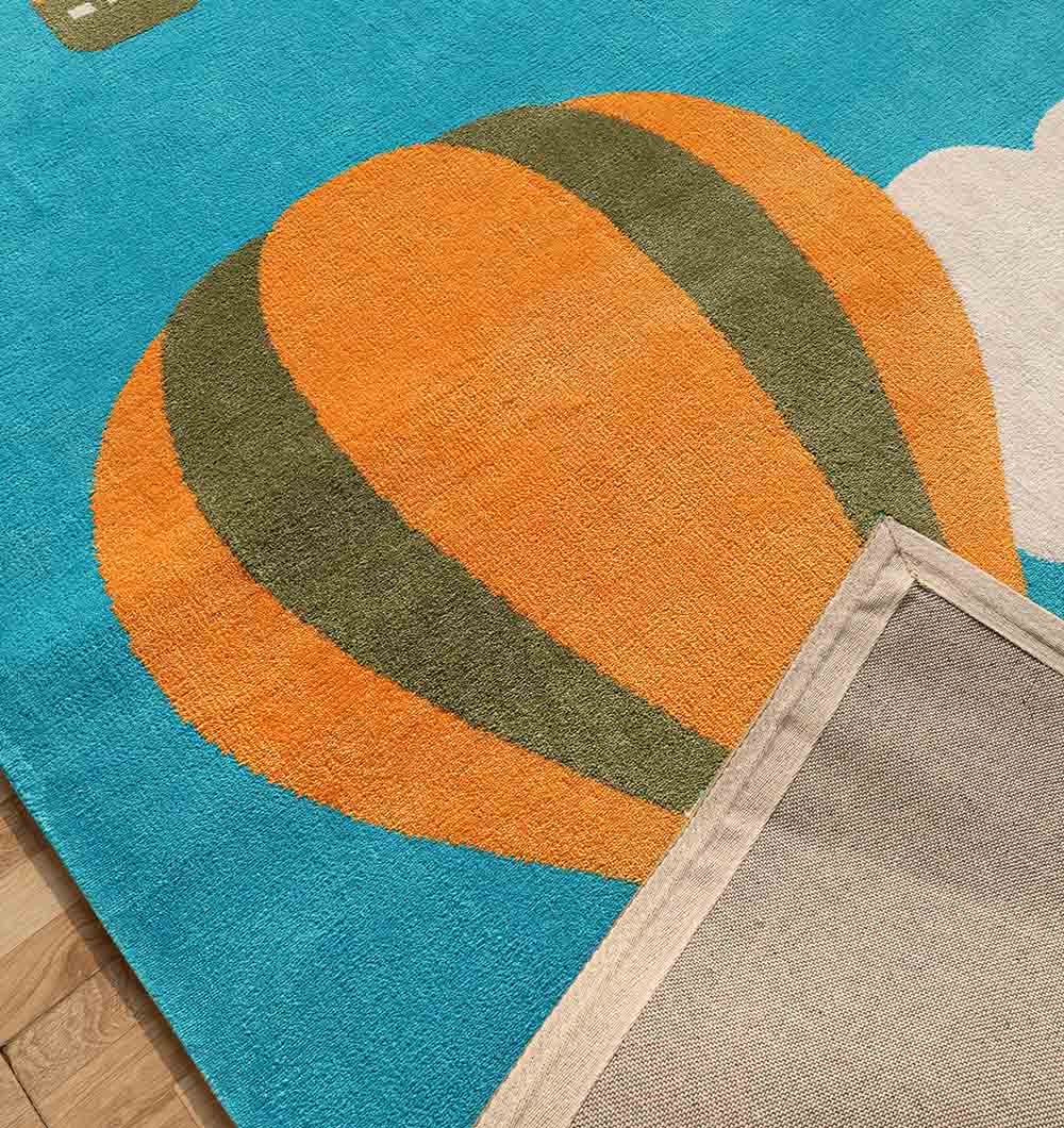 The RAMSHA HOME Modern Kids Area Rug - KD 012 (120x180 cm) features vibrant orange and green hot air balloons on a sky blue background, with a flipped corner displaying its beige backing.