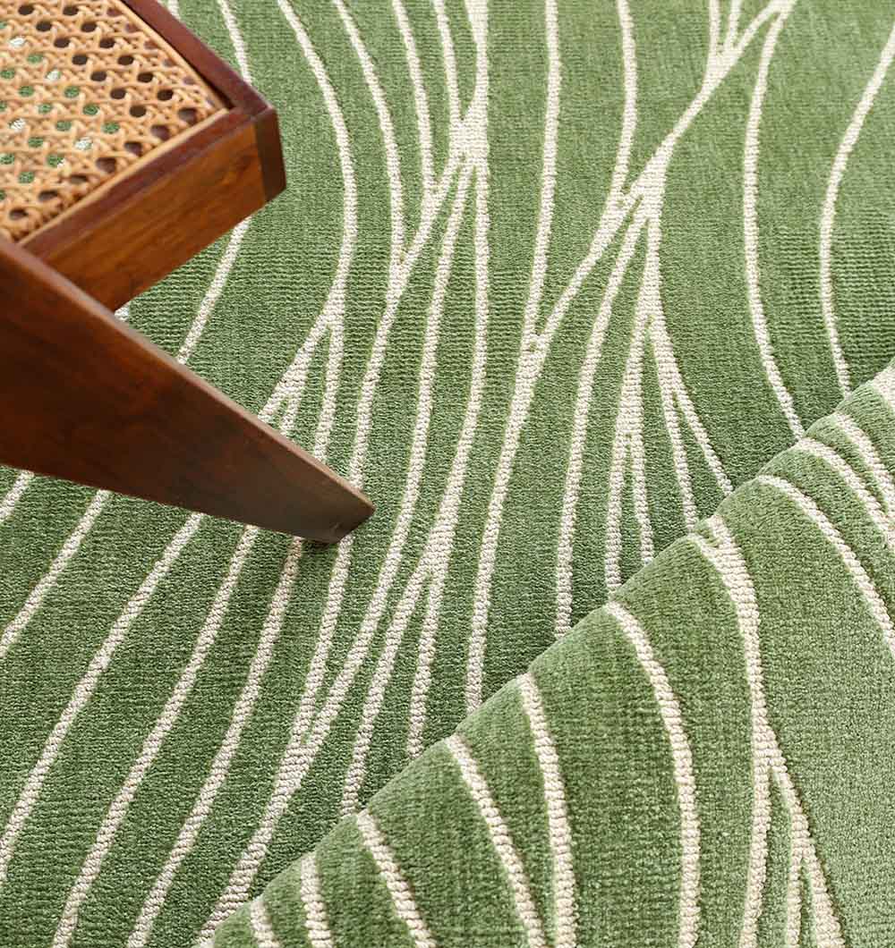 The RAMSHA HOME Leaf Vein Line Pattern Green & Ivory Rug – KD 015 is partially rolled, with the corner of a wooden chair featuring a woven cane seat visible in the upper left of the image.