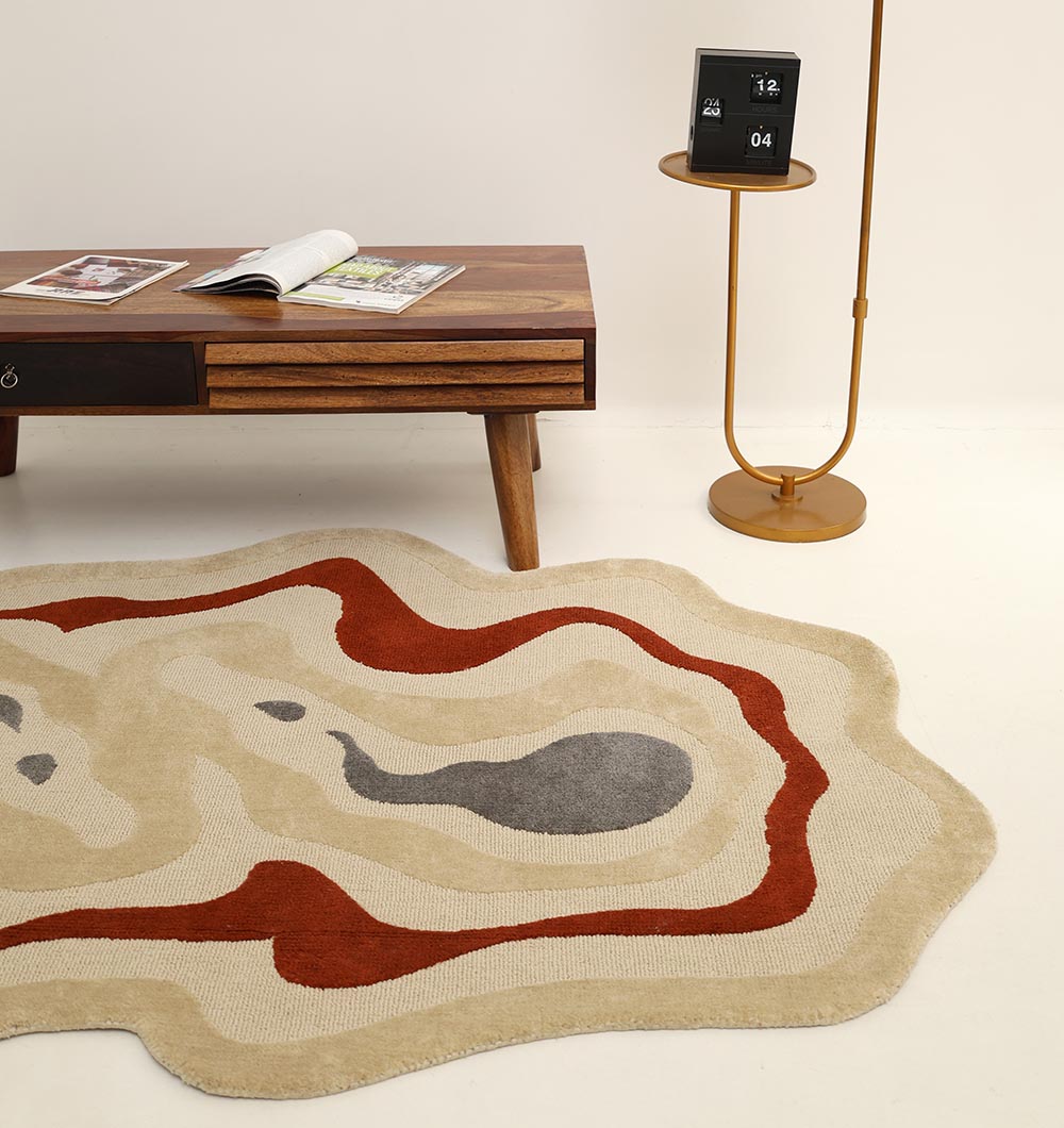 The RAMSHA HOME Amoeba Shaped Multi Color Rug (KD 016, 130x200 cm) in beige, red, and gray rests on a white floor beside a wooden coffee table with books and a round side table holding a calendar and digital clock.