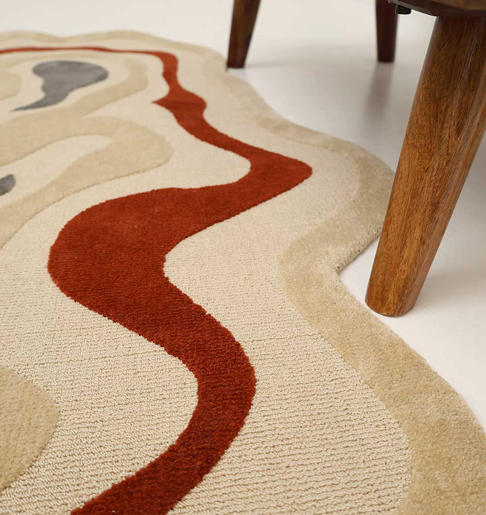 A close-up of the RAMSHA HOME Amoeba Shaped Multi Color Rug High Low Pile Rug 130x200 cm – KD 016, in beige with red and gray curves, is shown beside wooden furniture legs on a white floor.