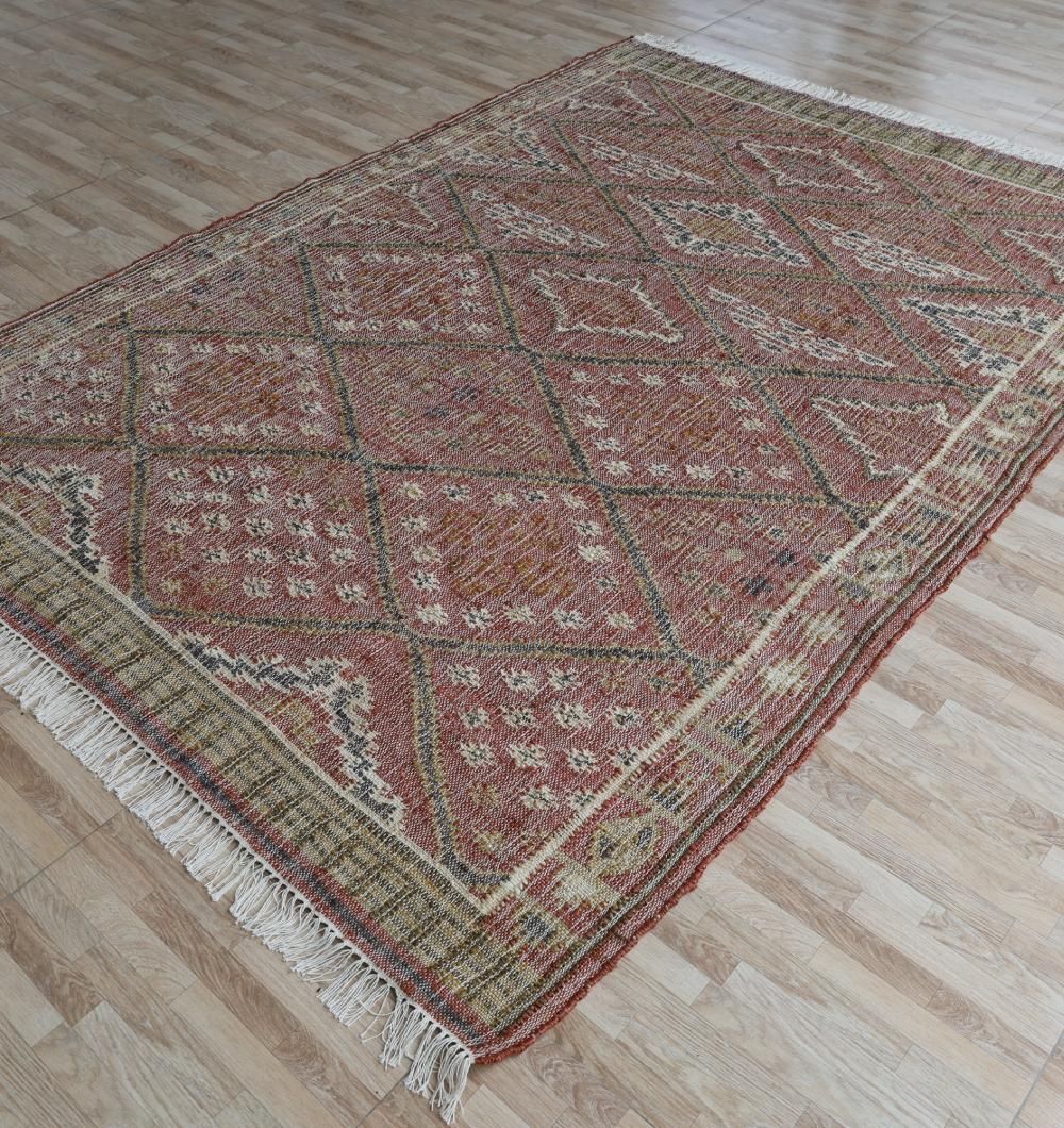 The RAMSHA HOME Durable Jute Kilim Flatweave Carpet – KLM 001 (200x300cm) features red, beige, and brown diamond and geometric patterns with natural texture and fringed edges, displayed on a light wood floor.