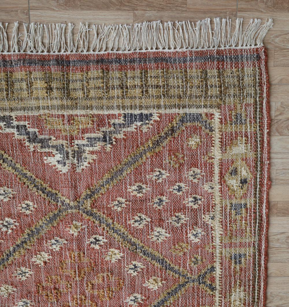 A close-up of the Durable Jute Kilim Flatweave Carpet – KLM 001 (200x300cm) by RAMSHA HOME shows its vintage-style geometric red, yellow, black, and cream pattern with white fringe, placed on a wooden floor highlighting its texture.
