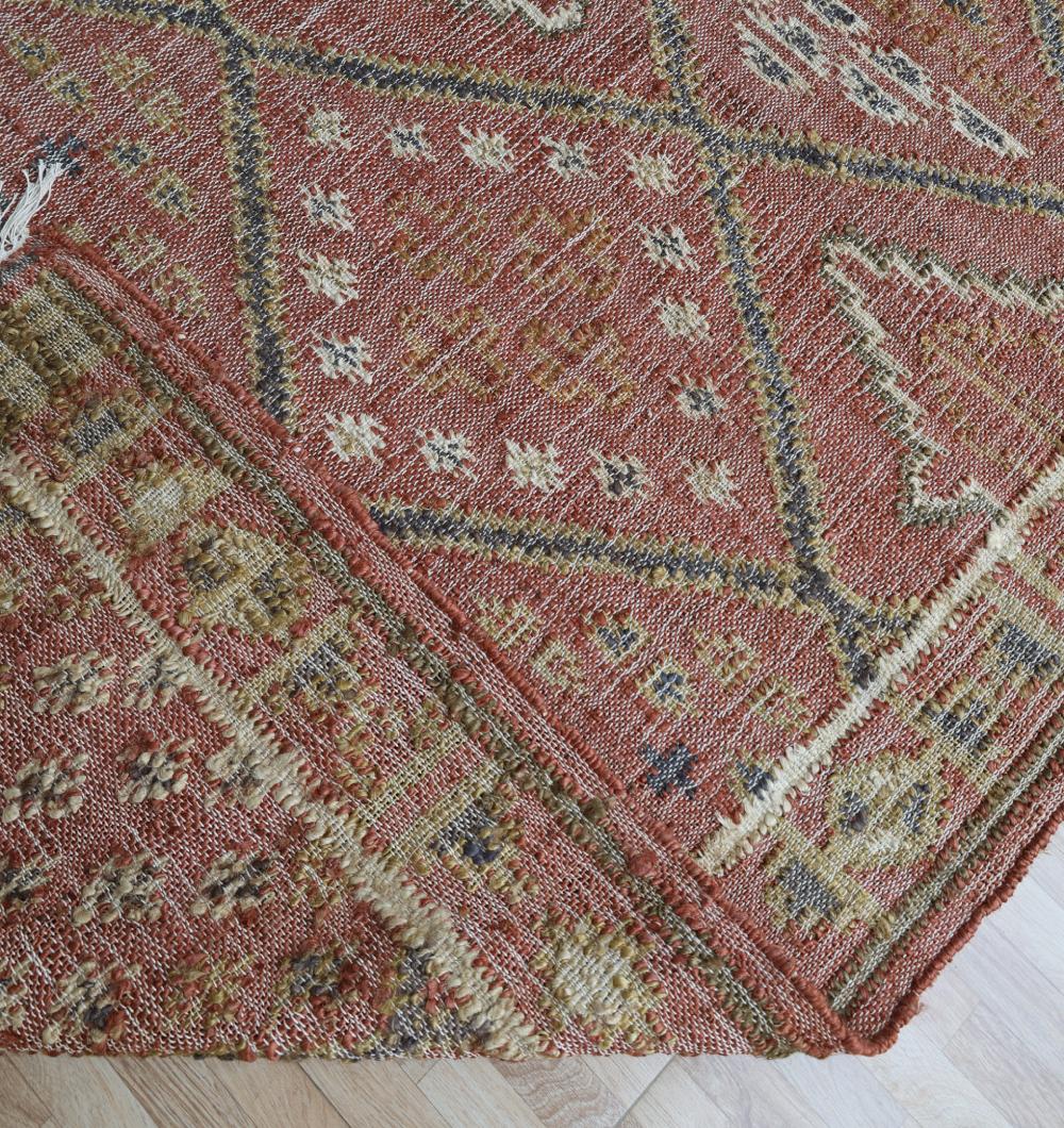 A close-up of the RAMSHA HOME Durable Jute Kilim Flatweave Carpet – KLM 001 (200x300cm), featuring geometric and diamond patterns in rust, beige, and yellow tones with natural texture, shown slightly folded on a light wooden floor.