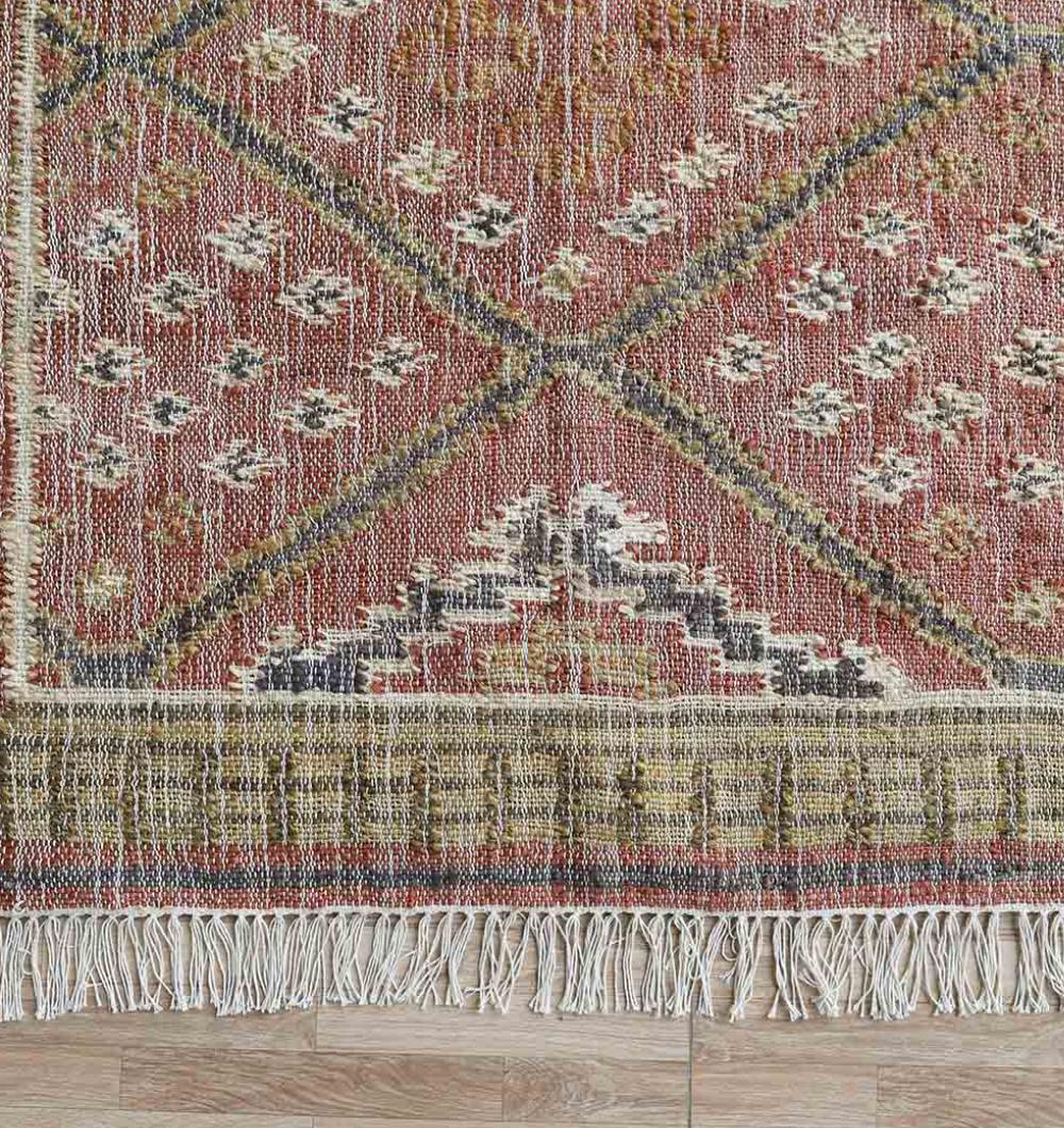 A close-up of the RAMSHA HOME Durable Jute Kilim Flatweave Carpet – KLM 001 (200x300cm) showcases its vintage geometric patterns in red, beige, black, and gold with a bottom fringe, placed on a light wooden floor for natural texture contrast.