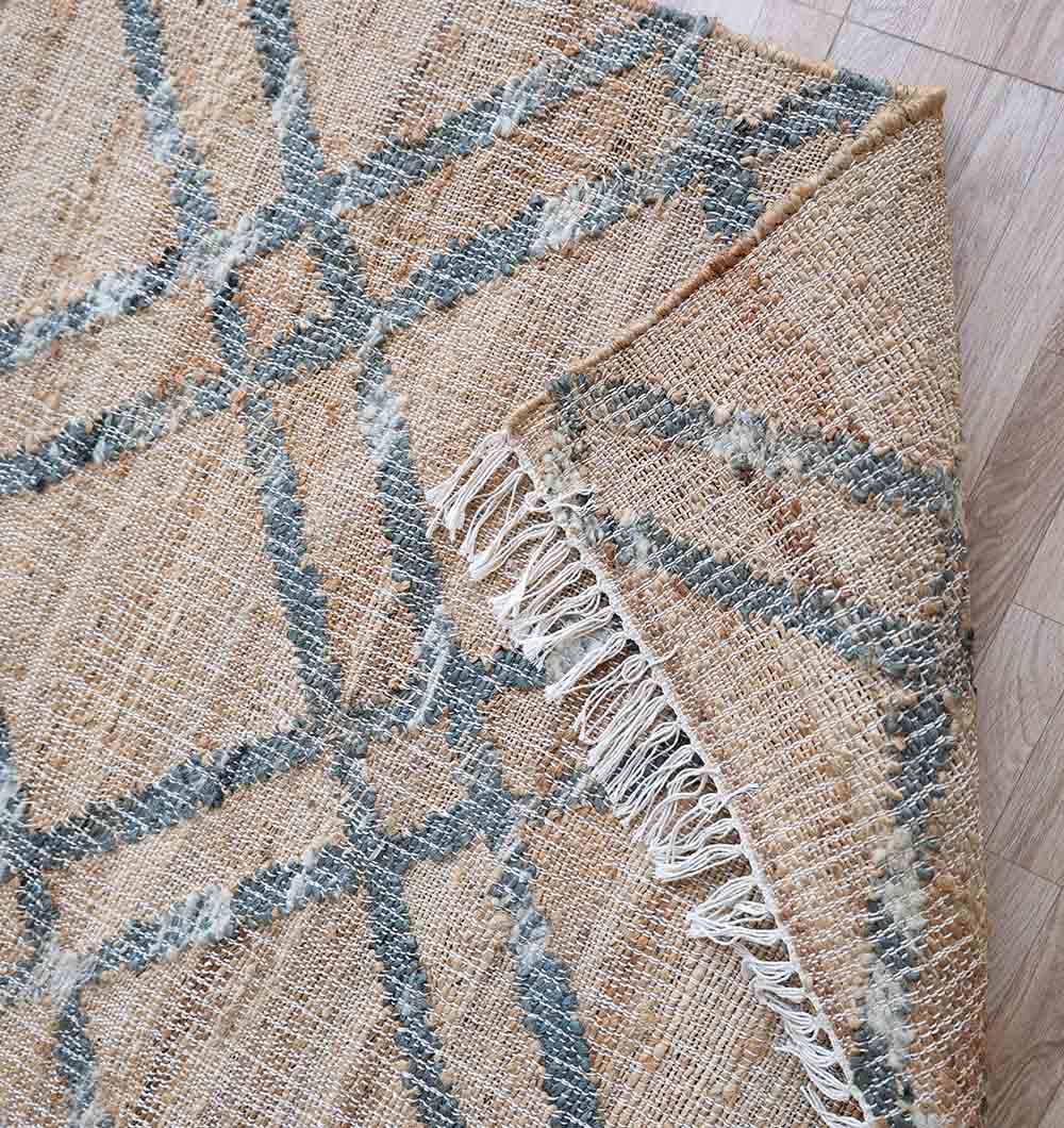 A close-up of the RAMSHA HOME Fringes Jute & Cotton Kilim Flatweave Carpet - KLM 002 (170x240cm) in beige and gray, partially flipped to show its fringe and texture, displayed on a light wooden floor.
