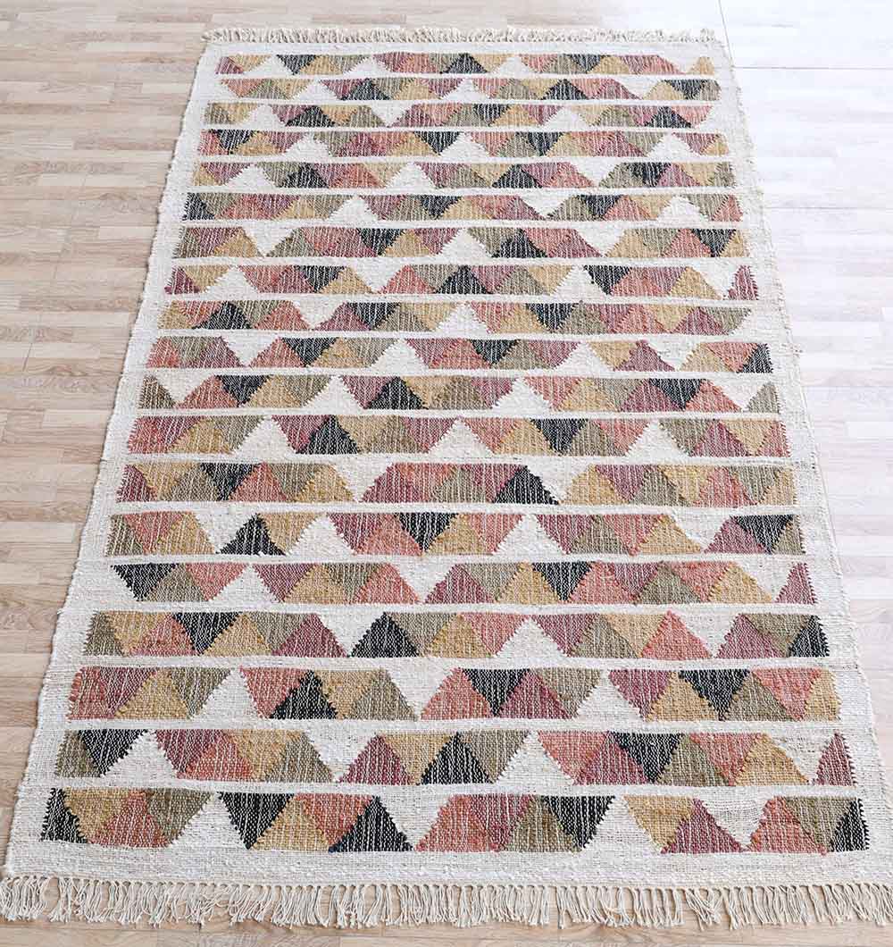 The RAMSHA HOME Geometric Jute Tribal Kilim Rug – KLM 003 (170x240cm) features colored triangles in black, brown, mustard, and rust on cream with fringed edges. Its natural texture adds warmth to light wood floors.