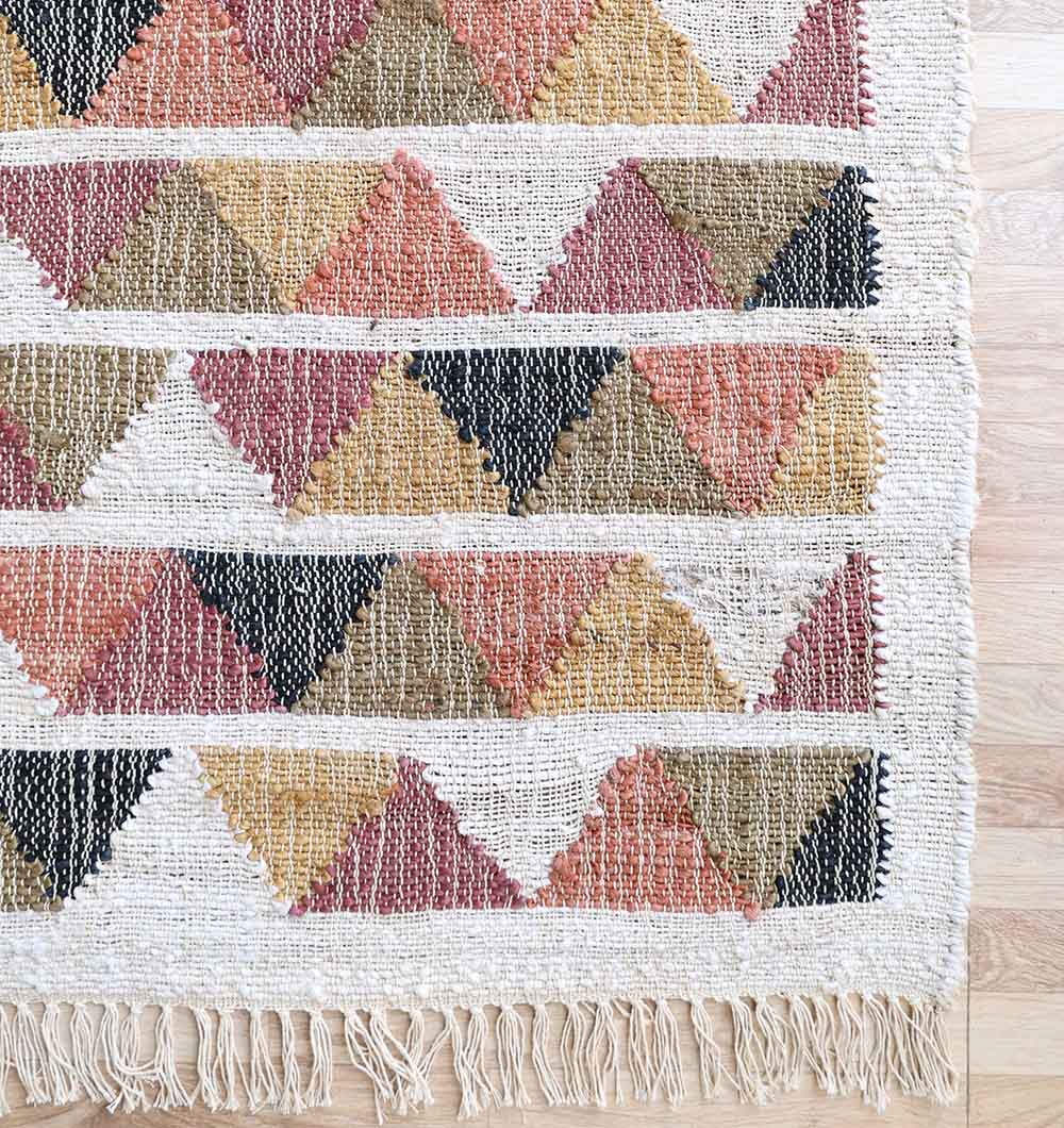 The RAMSHA HOME Geometric Jute Tribal Kilim Rug – KLM 003 (170x240cm) features rows of orange, mustard yellow, black, and brown diamonds on a cream background with natural texture and fringed edges, shown atop a wood floor.