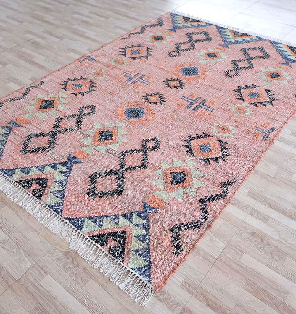 The RAMSHA HOME Handwoven Jute Flatweave Pink Kilim Rug – KLM 005 (170x240cm) features geometric patterns in pink on a light wood floor, with fringed edges on the shorter sides for added texture and style.