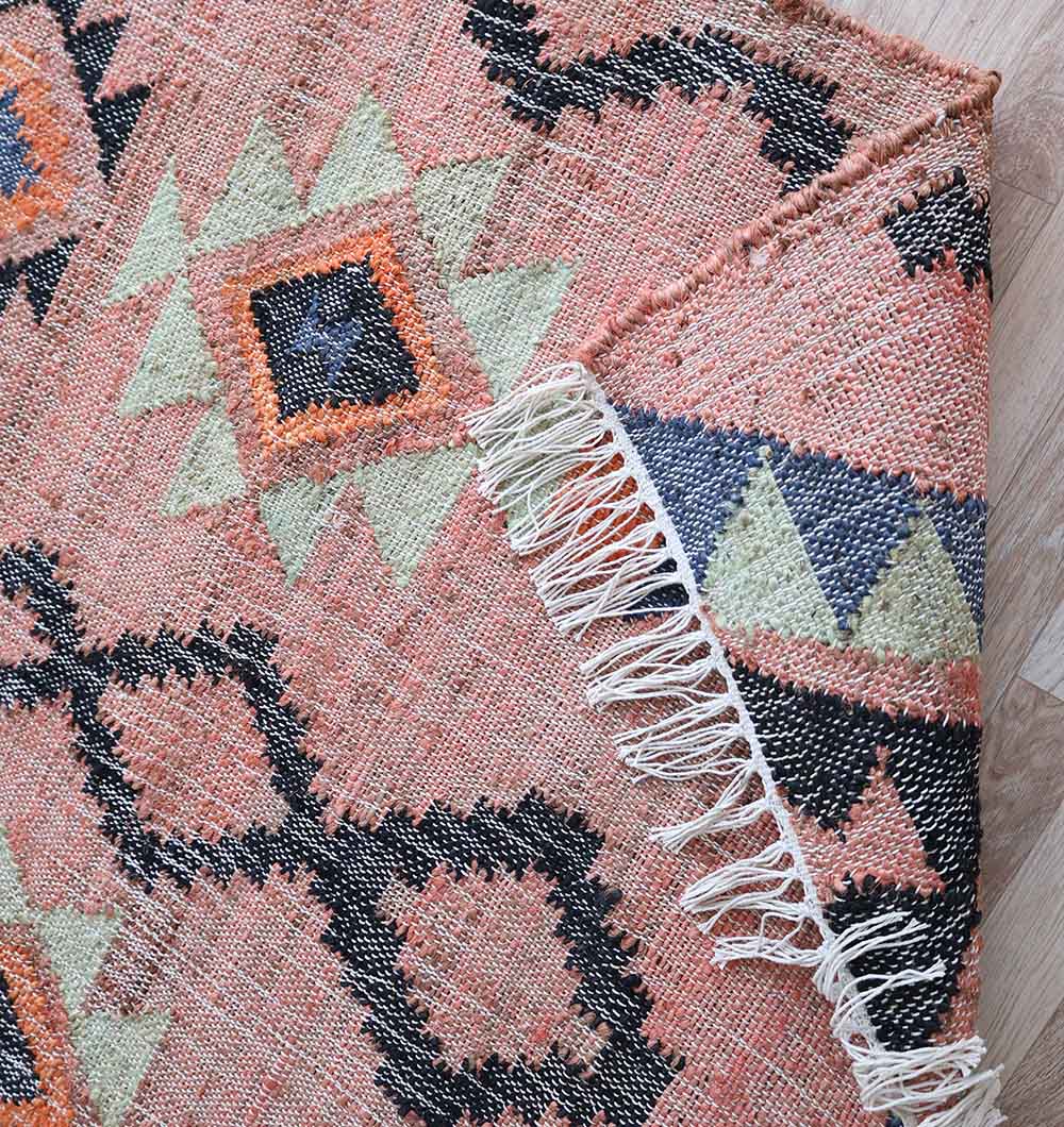The RAMSHA HOME Handwoven Jute Flatweave Pink Kilim Rug – KLM 005 (170x240cm) features geometric diamond patterns in pink, black, cream, orange, and green with fringed edges, displayed on a light wooden floor.