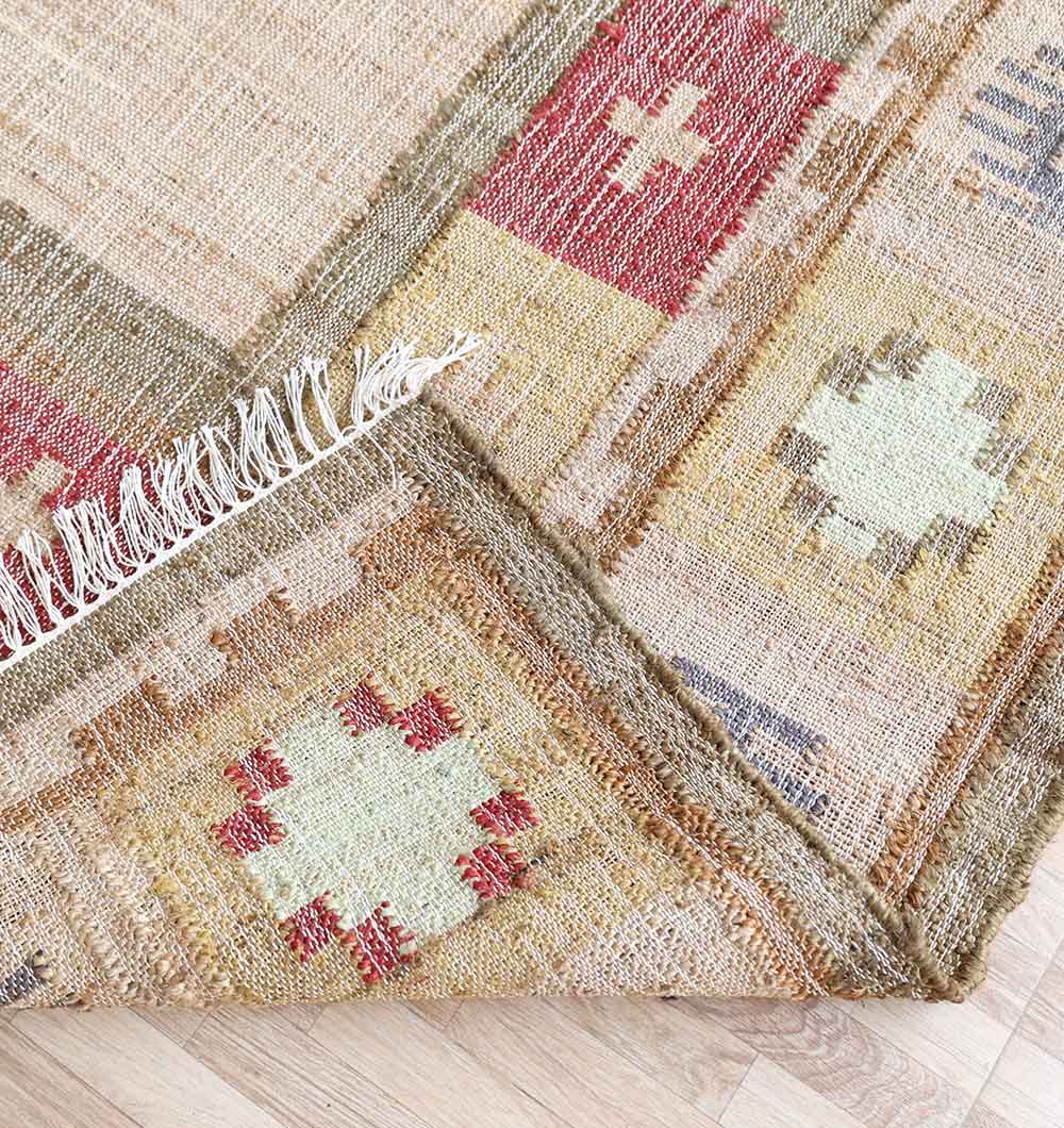 A close-up of the RAMSHA HOME Symmetrical Bordered Traditional Kilim Rug - KLM 007 (170x240cm) shows its earthy red, yellow, and cream geometric patterns. One corner is folded, displaying fringed edges and the wooden floor underneath.
