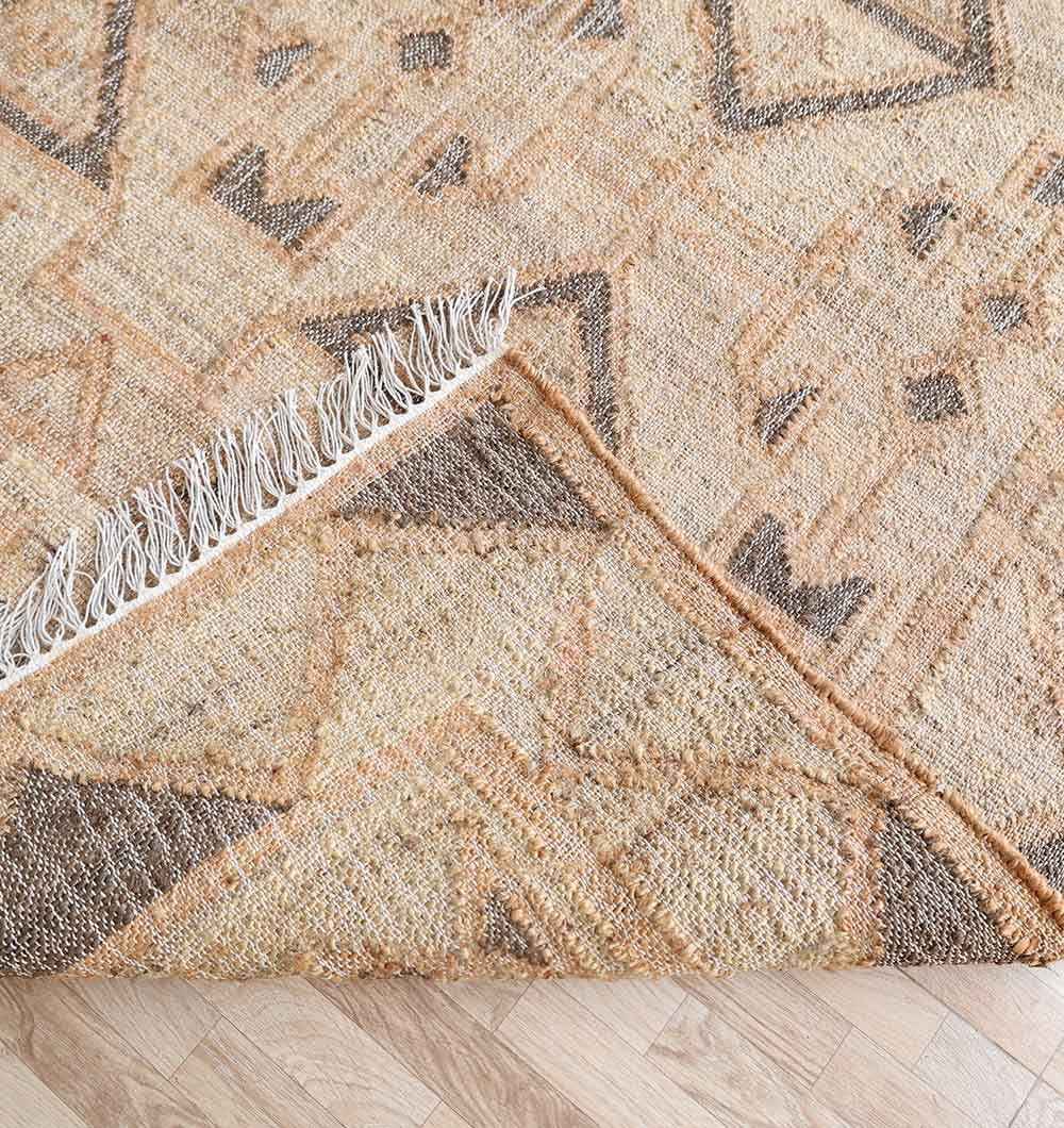 A close-up of the Minimalist Neutral Jute Tribal Kilim Rug - KLM 008 (170x240cm) by RAMSHA HOME, showing its brown and tan geometric pattern, folded corner, frayed edges, and intricate tribal design.