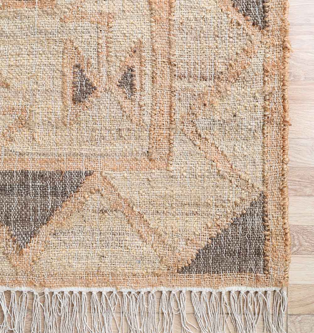 A close-up of the RAMSHA HOME Minimalist Neutral Jute Tribal Kilim Rug - KLM 008 (170x240cm) showing its beige and brown geometric pattern and fringed edges, placed on a wooden floor.