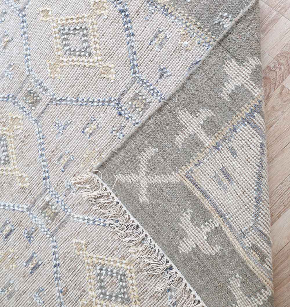 A close-up of the RAMSHA HOME Neutral Diamond Geometric Flatweave Kilim Rug - KLM 009 (170x240cm), in grey with beige and light blue geometric patterns, shows its fringed edge and underside as it rests on a wooden floor.