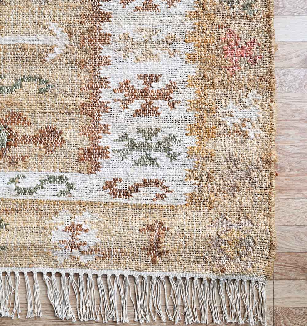 A close-up of the RAMSHA HOME Jute Cotton Blend Turkish Styled Kilim Rug - KLM 011 shows beige, brown, green, and rust geometric patterns, natural texture, fringed edges, and a wood floor underneath.
