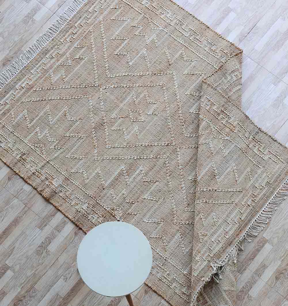 A beige RAMSHA HOME Jute Cotton Blend Kilim Rug (KLM 012, 170 x 240 cm) with geometric patterns is partially folded on a light wood floor, with a round white side table nearby.