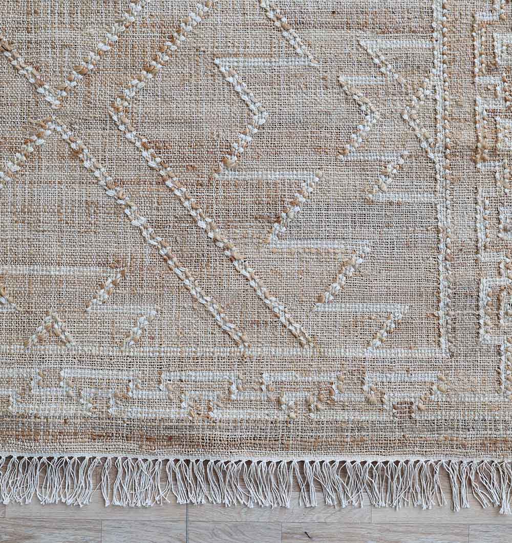 A close-up of the RAMSHA HOME Jute Cotton Blend Kilim Rug (KLM 012, 170 x 240 cm), featuring geometric patterns in beige and cream with fringed edges, displayed on a light wooden floor.