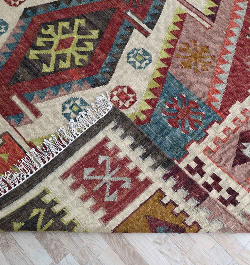 The RAMSHA HOME Authentic Turkish Geometric Kilim Rug - KLM 013, inspired by traditional tribal designs, showcases red, blue, yellow, green, and cream shapes with fringed edges lying on a light wood floor.