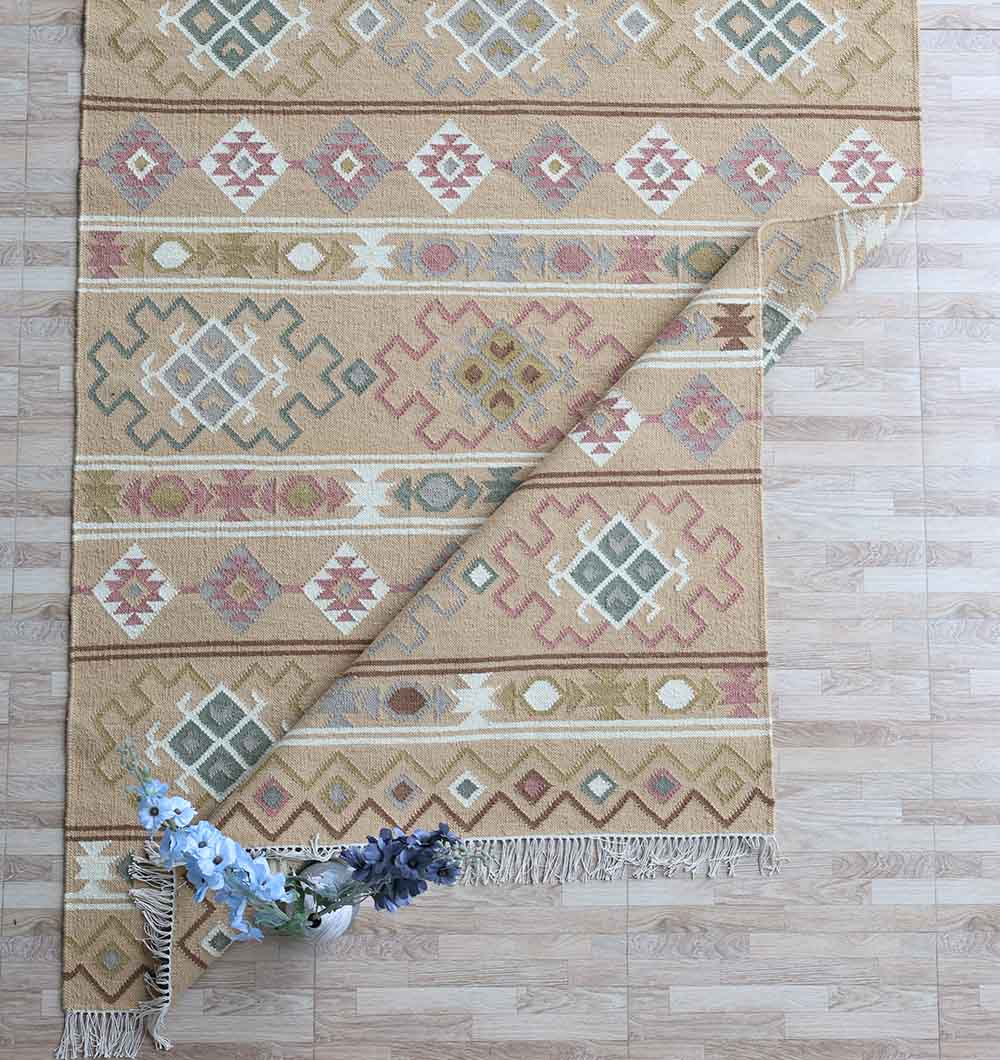 The Handwoven Neutral Beige Wool Kilim Rug (KLM 014) from RAMSHA HOME, in muted pink, green, and gold geometric patterns, is partly rolled on a light wood floor with a vase of blue and purple flowers at its fringed edge.