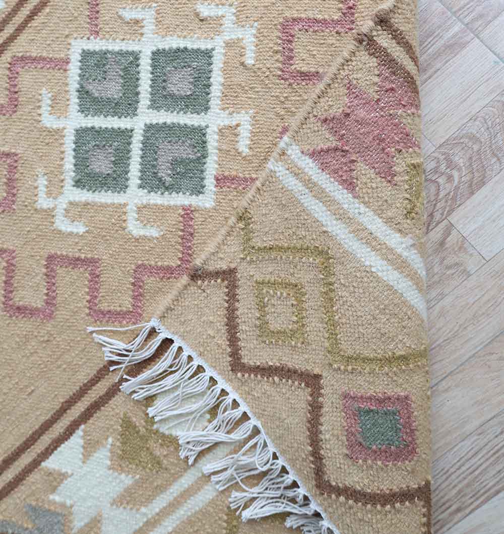 A close-up of the RAMSHA HOME Handwoven Neutral Beige Wool Kilim Rug - KLM 014 shows its geometric pattern in beige, pink, green, and white, with fringe and underside visible on a light wooden floor.