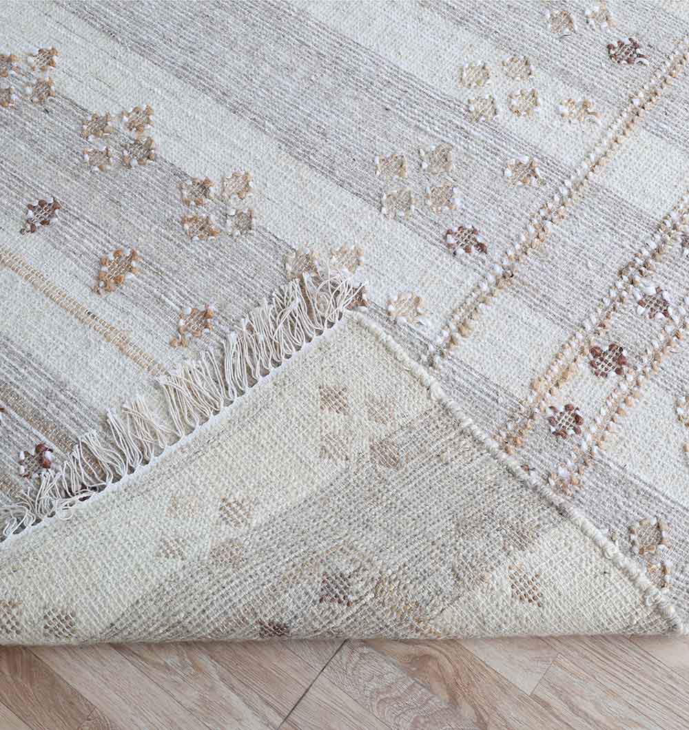 A close-up of the RAMSHA HOME Handwoven Boho Area Rug Grey Kilim Rug (KLM-016, 170x240cm), featuring textured grey flatweave with subtle geometric patterns and fringe, partially folded on a light wood floor.