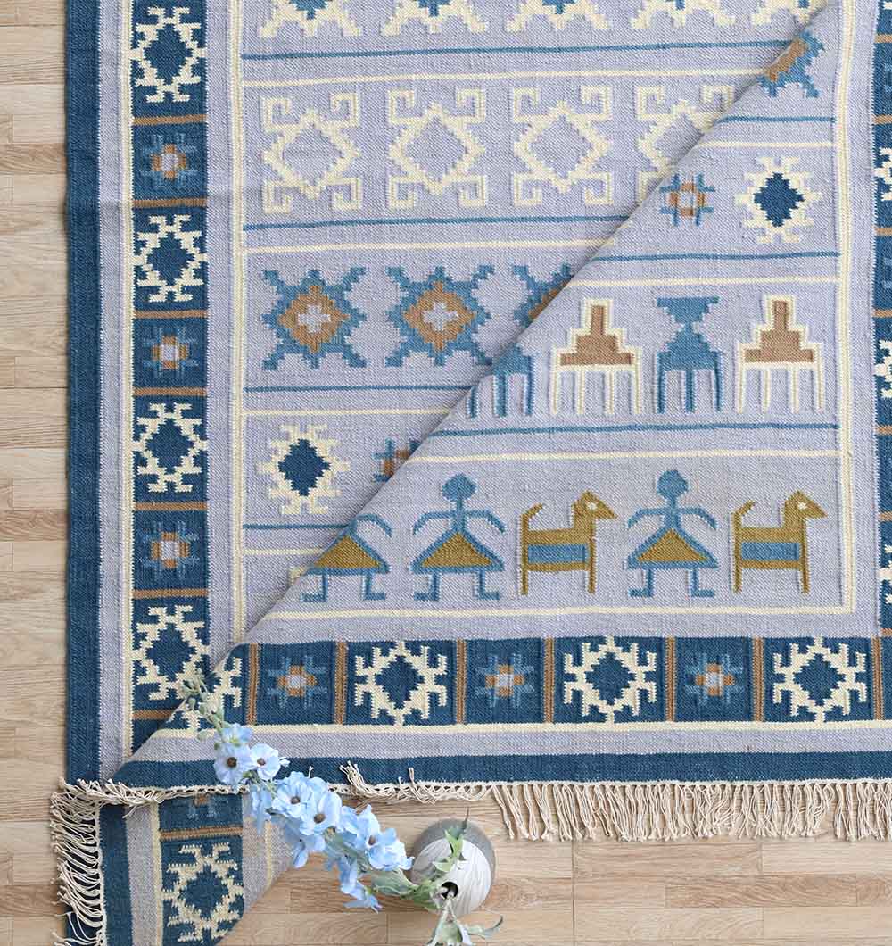 The RAMSHA HOME Kilim Rug – Blue Multicolor Tribal Flatweave Carpet KLM-018, featuring geometric and animal patterns in blue, beige, and yellow, is partially folded on a wooden floor beside light blue flowers.