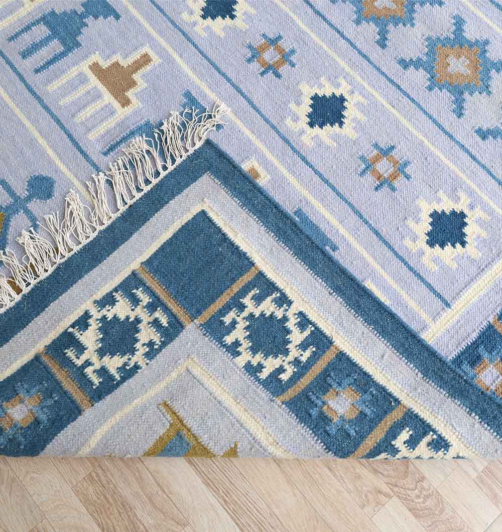 The RAMSHA HOME Kilim Rug – Blue Multicolor Tribal Flatweave Carpet KLM-018, featuring geometric motifs and fringed edges, is shown partially folded to reveal the underside on a light wooden floor.