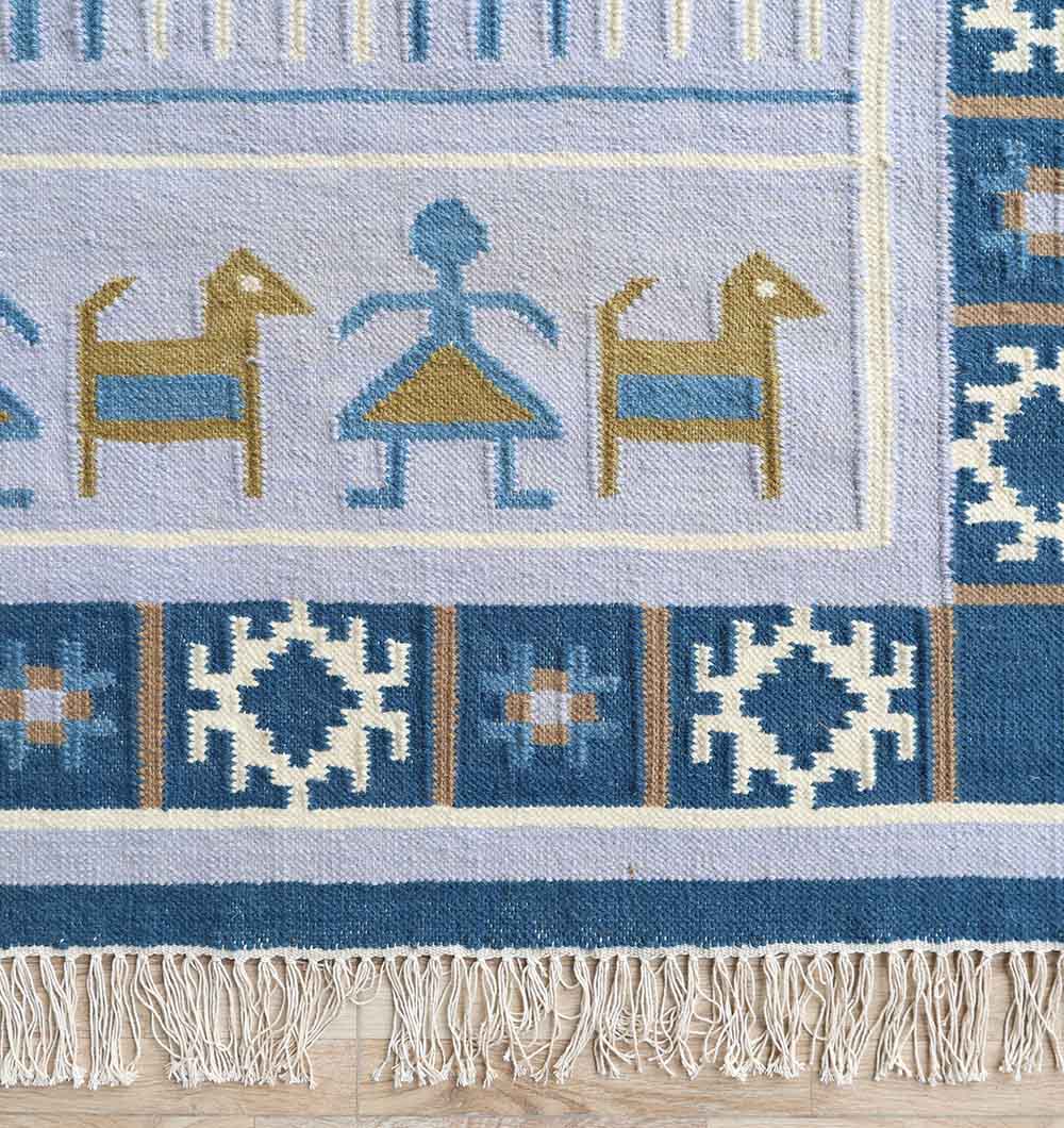 The RAMSHA HOME Kilim Rug – Blue Multicolor Tribal Flatweave Carpet KLM-018 features geometric motifs, stylized animals, a human figure, and a decorative blue, gold, and cream border on a light lavender background.