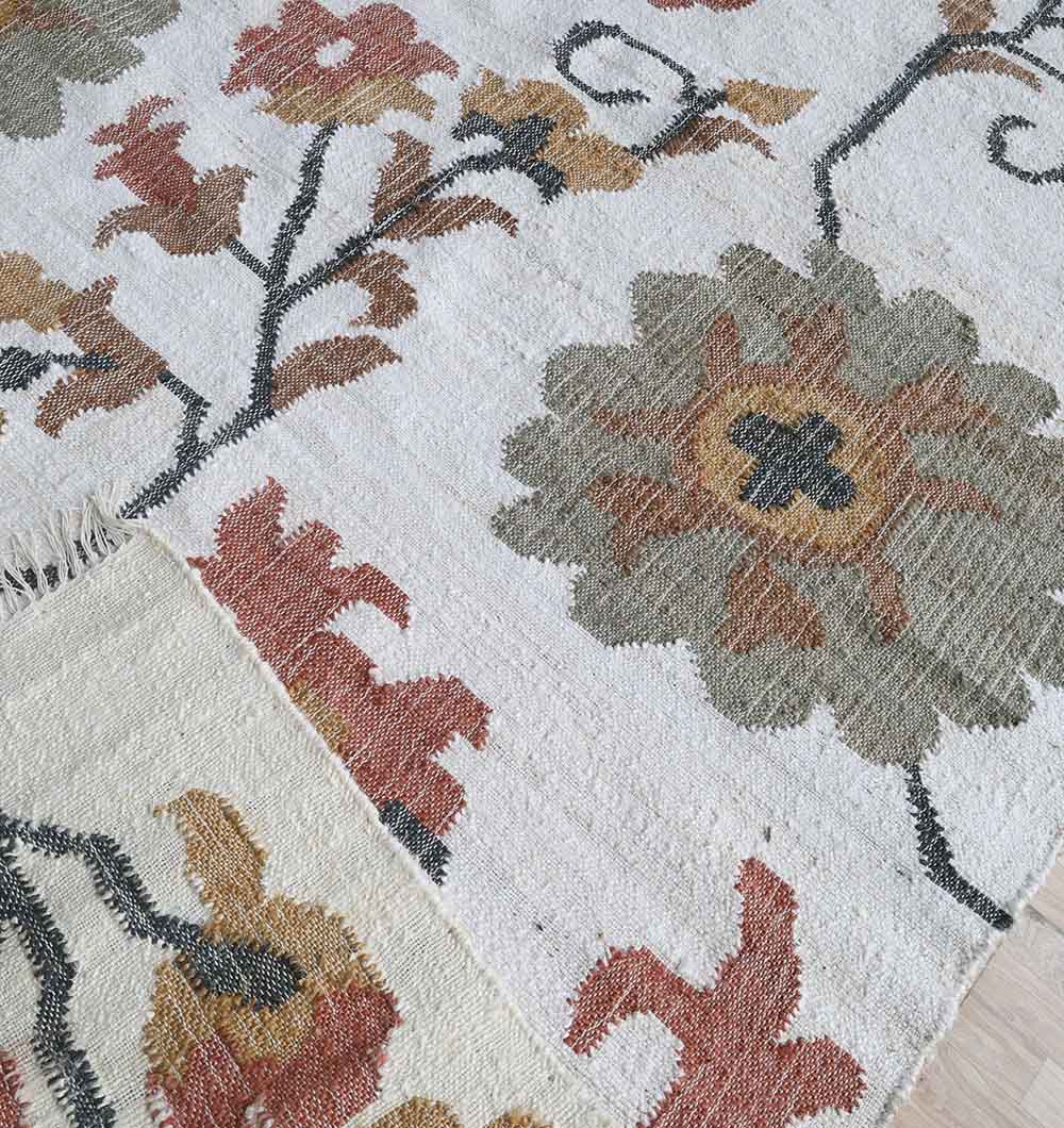 A close-up of the RAMSHA HOME Kilim Rug – Multicolor Floral Handmade Flatweave Carpet KLM-021 shows its green, orange, and red floral design on cream. A folded corner reveals the natural jute texture and detailed reverse side of this artisan piece.